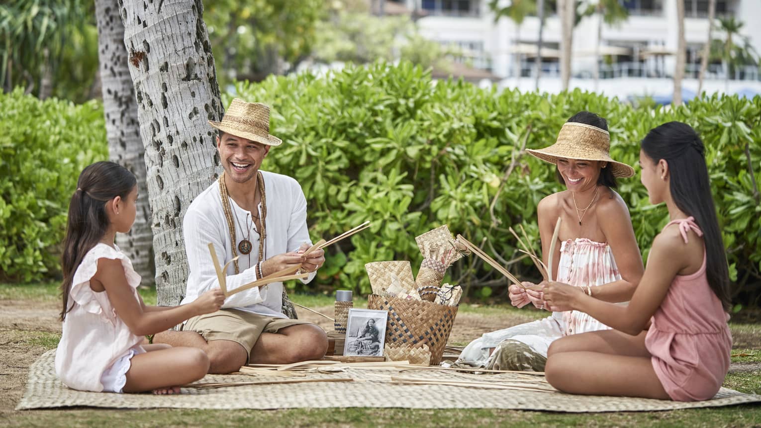 Two adults and two children sit on a blanket under a tree as they practice weaving