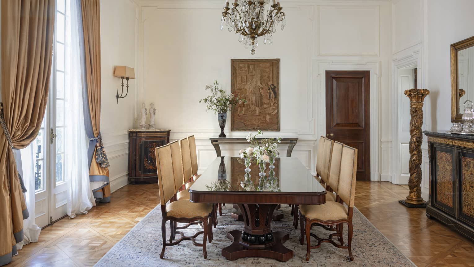 Suite dining room with rectangular table set with eight high-back chairs, a crystal chandelier overhead and tall floor-to-ceiling French doors on one side draped with gold curtains