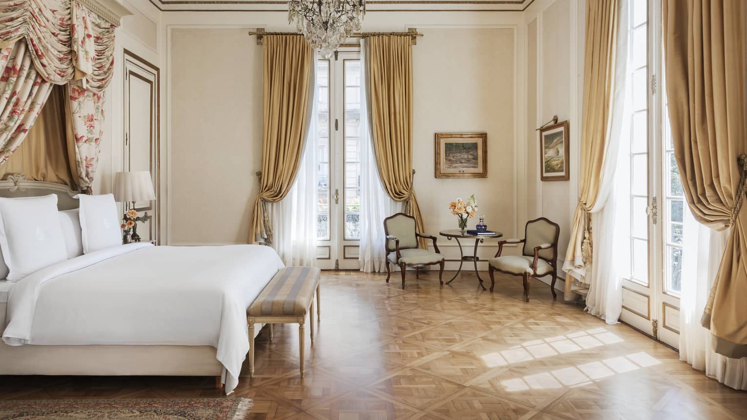 Suite bedroom with parquet flooring, a crystal chandelier, large white bed and two sets of floor-to-ceiling French doors