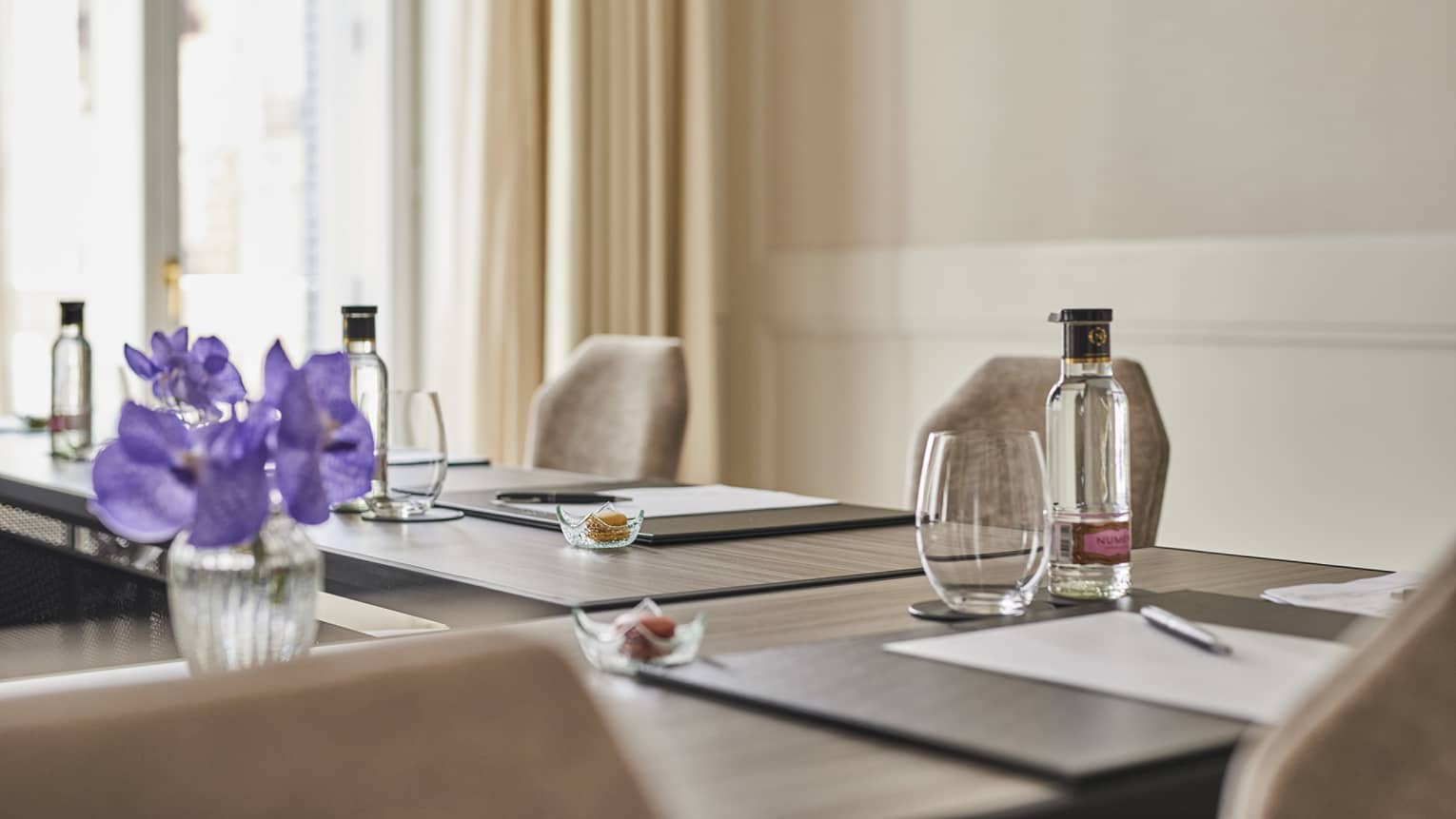 A meeting room with beige walls and curtains, a large window, chairs, and brown tables set with paper, pens, drinking glasses, clear bottles and tiny bowls, plus a vase of purple flowers