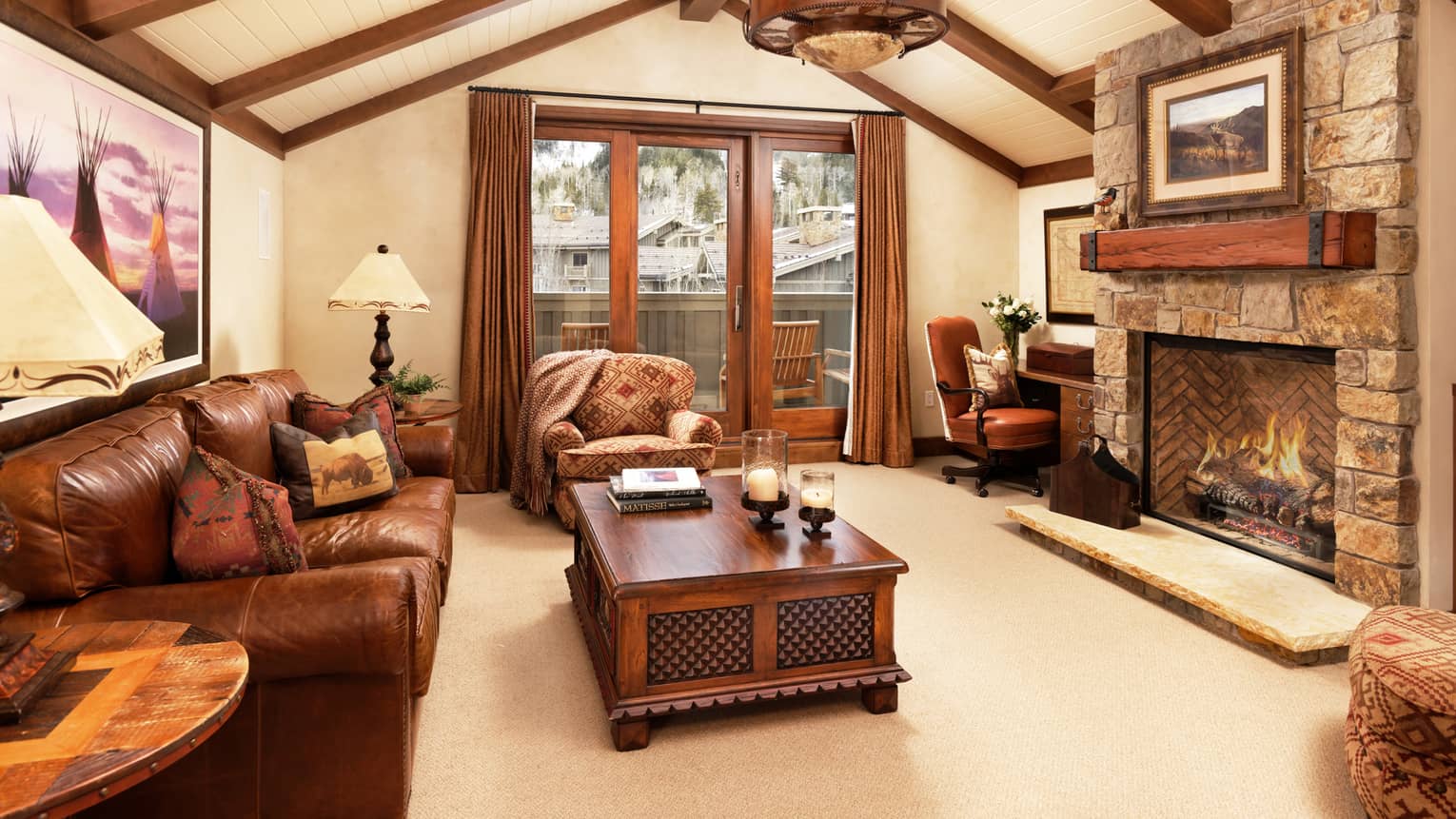 Rustic living room with leather sofa, armchair, tables, desk, stone fireplace, vaulted wood-beam ceiling and balcony doors with mountain view