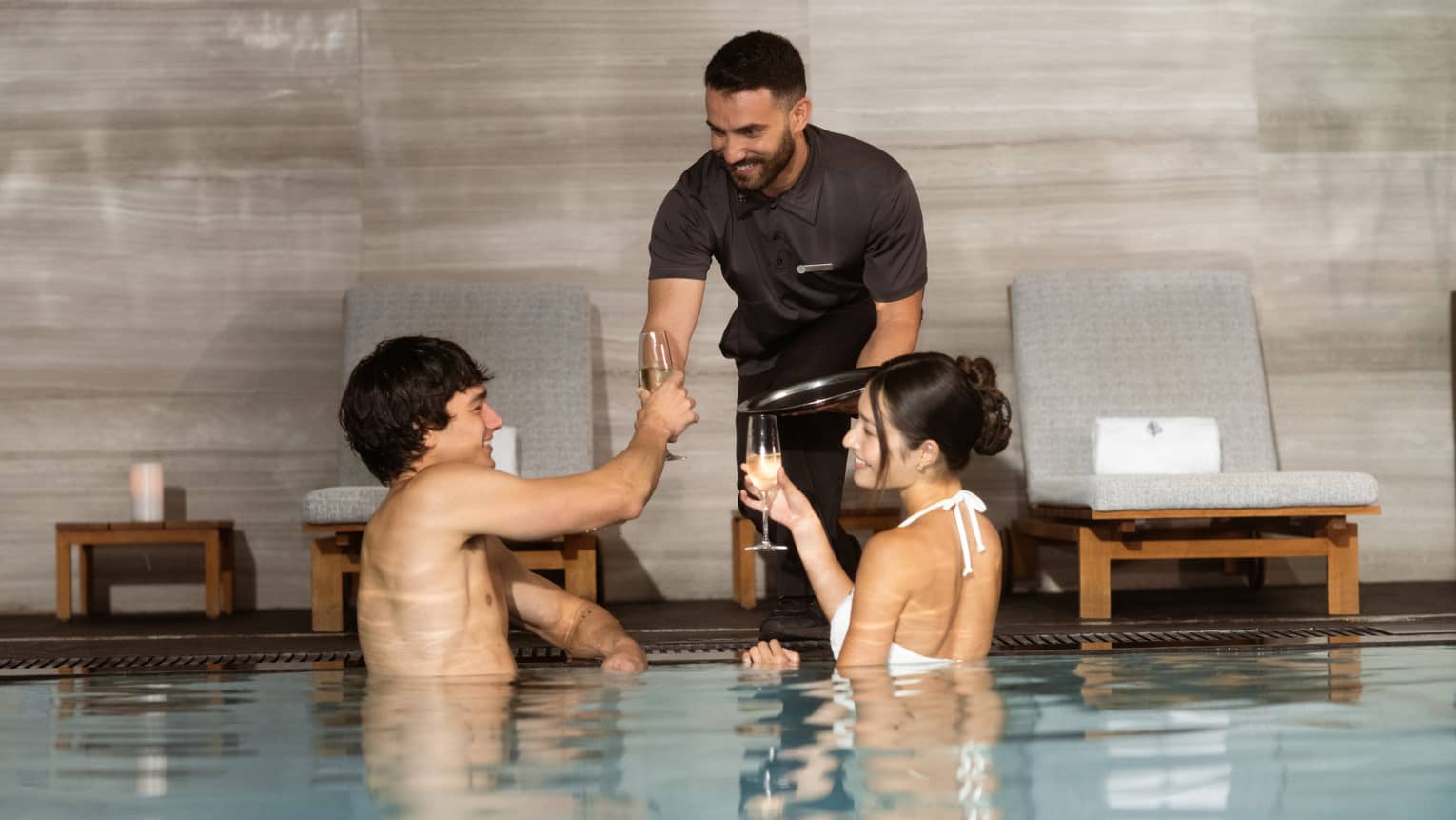 Two smiling guests in an indoor pool with glassy water and grey marble walls are served drinks in champagne flutes.