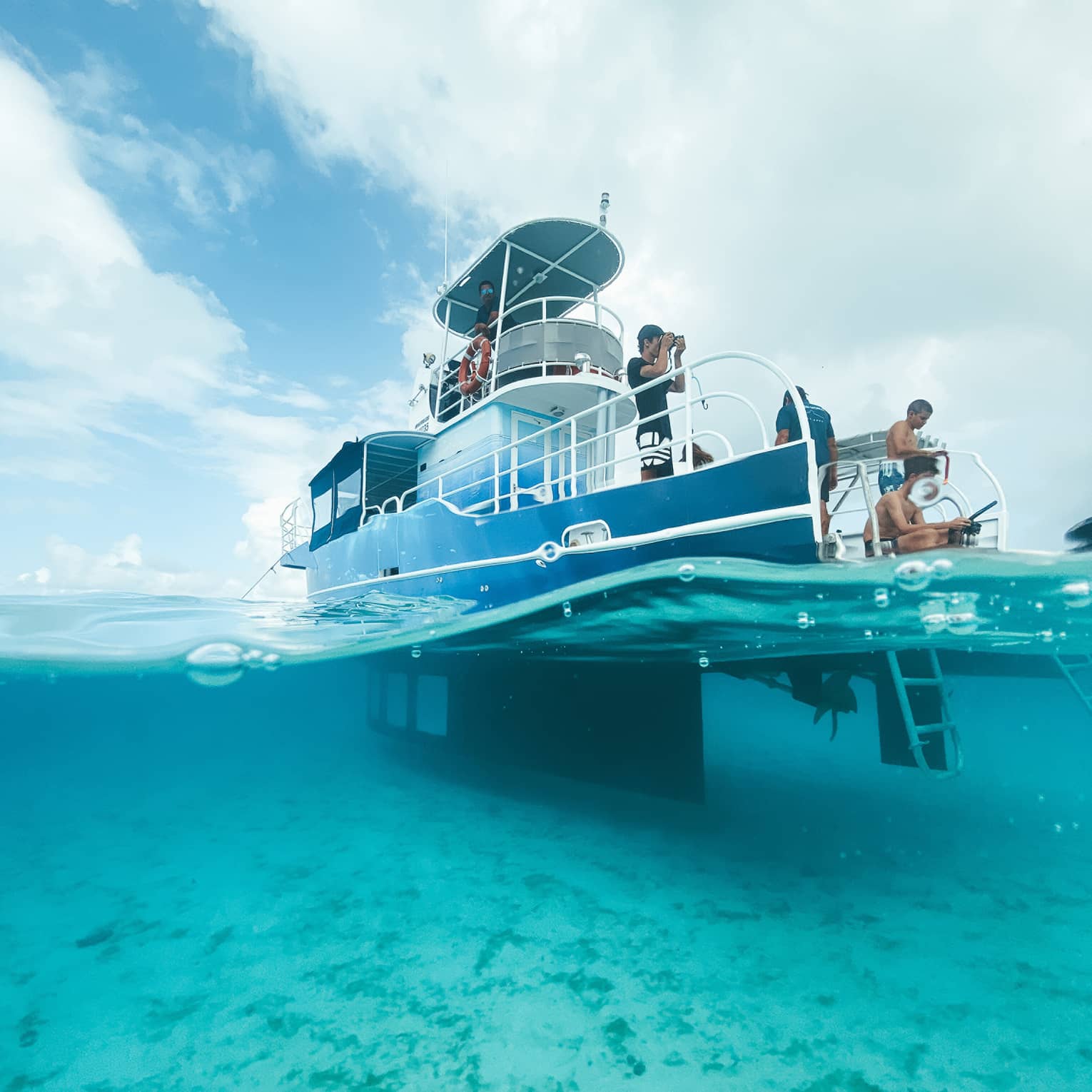A boat with an underwater viewing cabin anchored in the ocean; above water, guests relax and take photos from the deck.