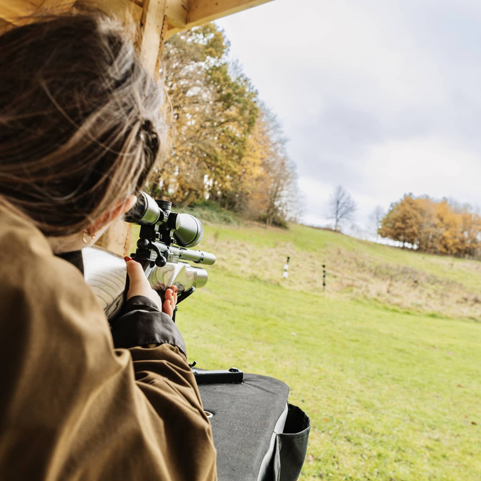Rear view of a person in a brown jacket aiming an air rifle at small, distant targets in a large field amid autumn trees.