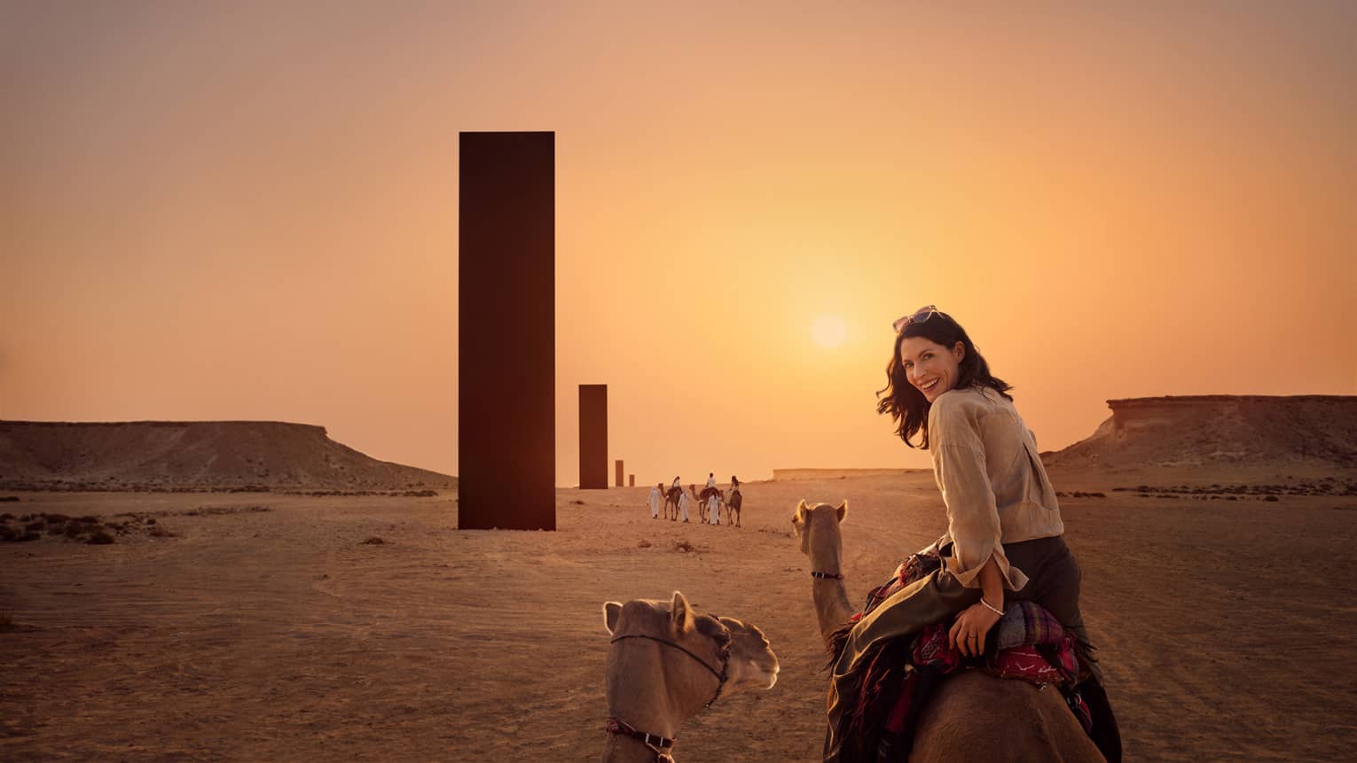 Person riding a camel looks over their shoulder and smiles with a row of black monoliths rising out of the desert landscape in front of them, all lit by an orange glow from the sunset