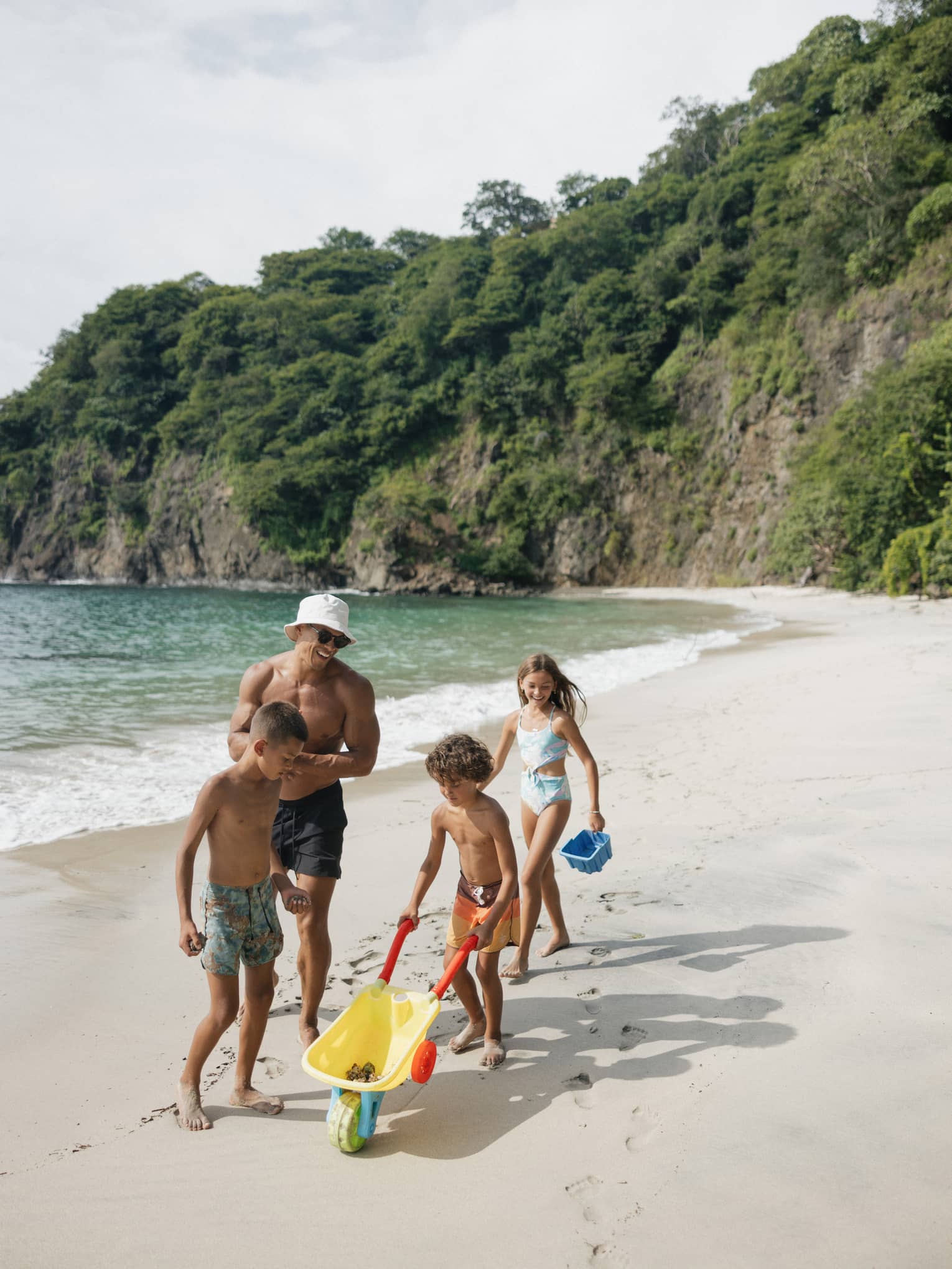 A parent and three young children play on a beach with a toy wheelbarrow and sand toys