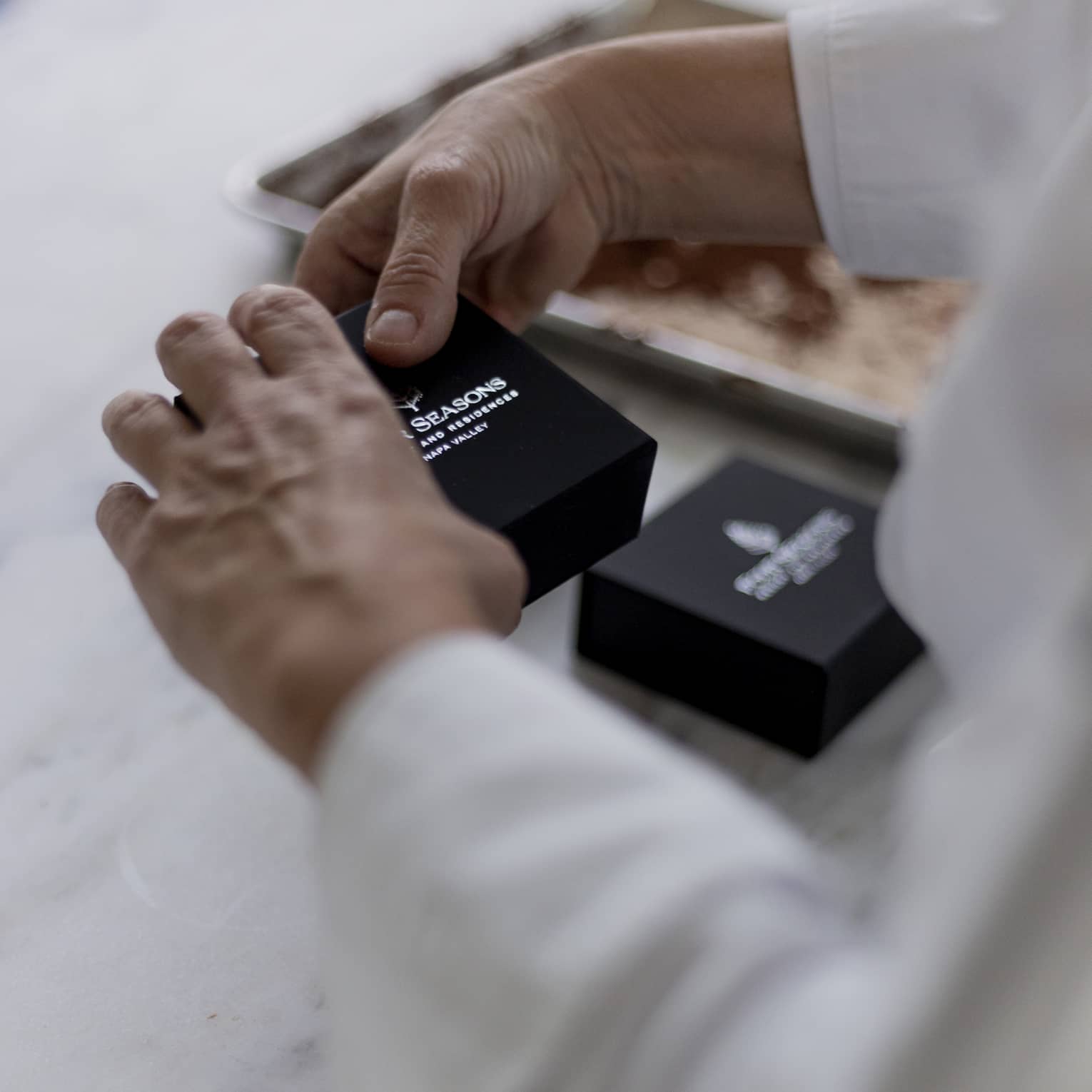 Hands holding a small, black box with the Four Seasons name and logo in silver; a blurred cookie sheet in the background.