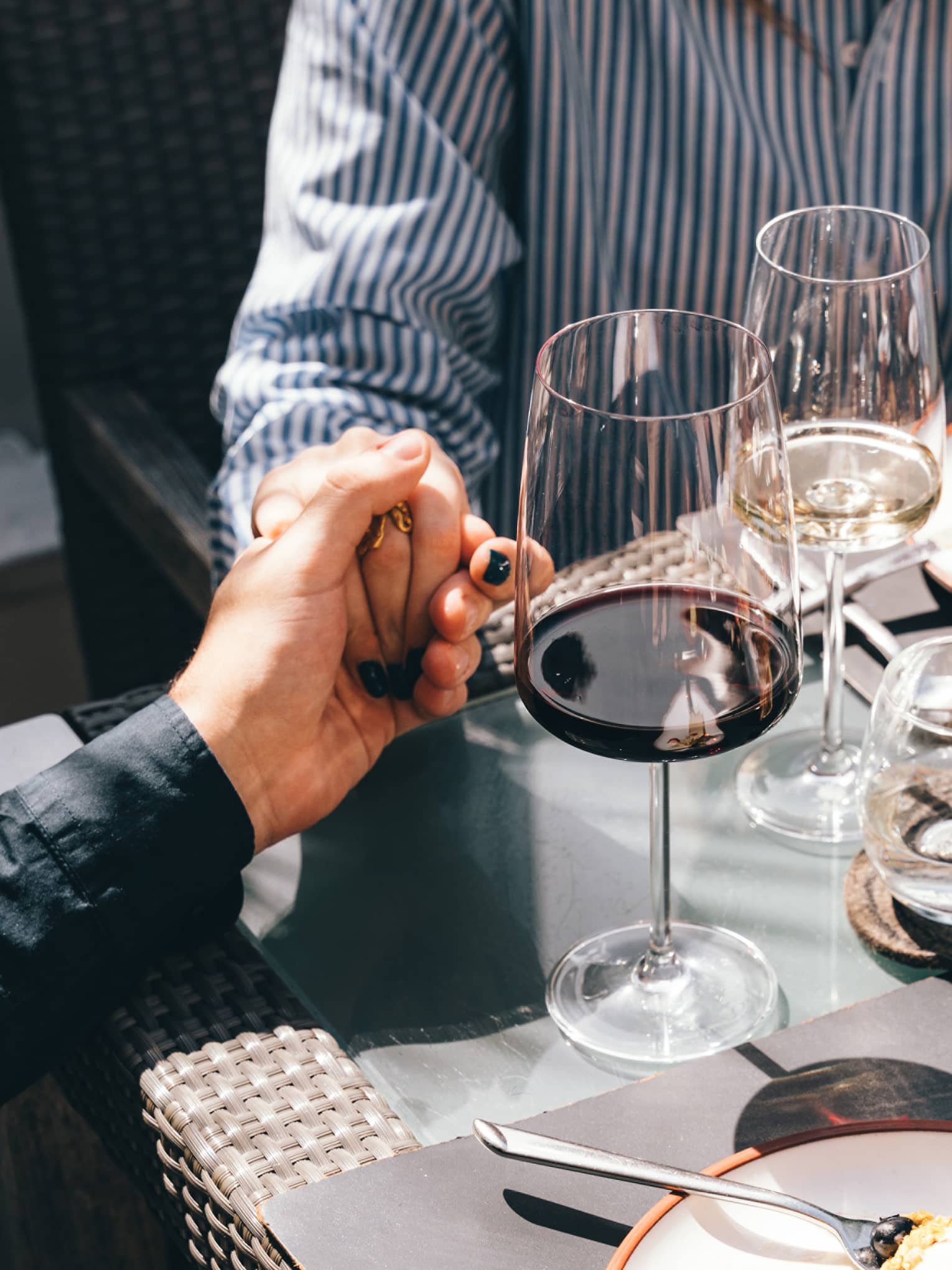 Close up of a couple holding hands as they're seated across from each other at an outdoor table, with a glass of red wine and a glass of white wine set before them