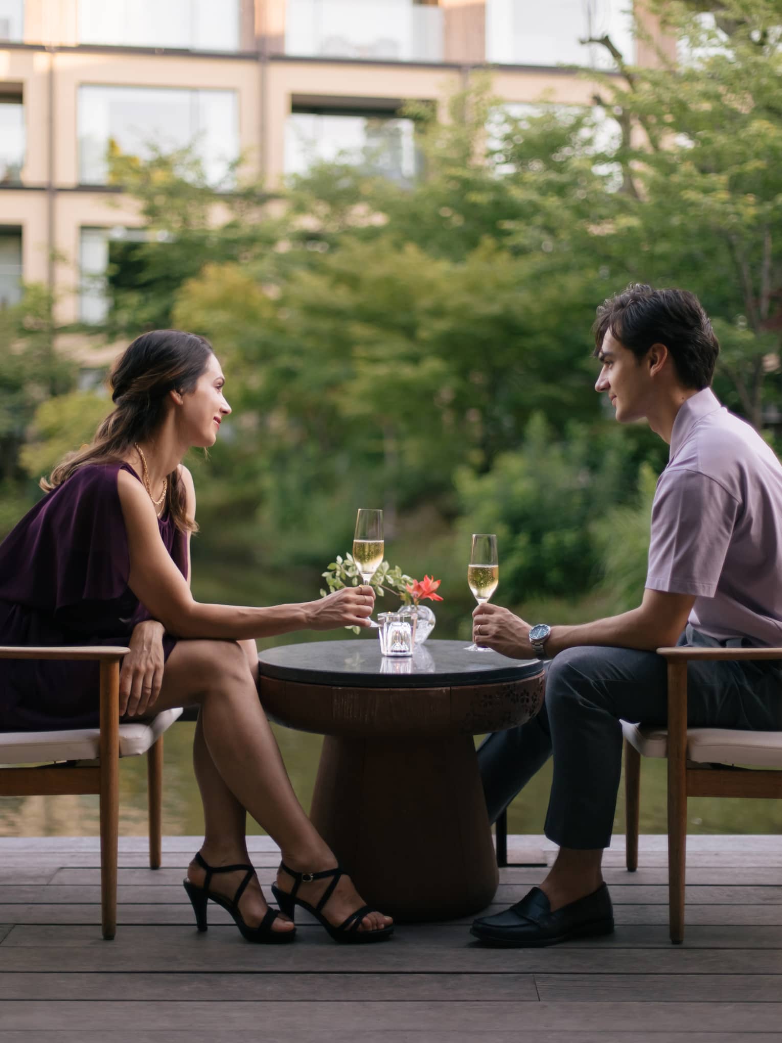 Profile view of couple at a table gazing at each other, champagne glass in hand, lush greenery in the background.