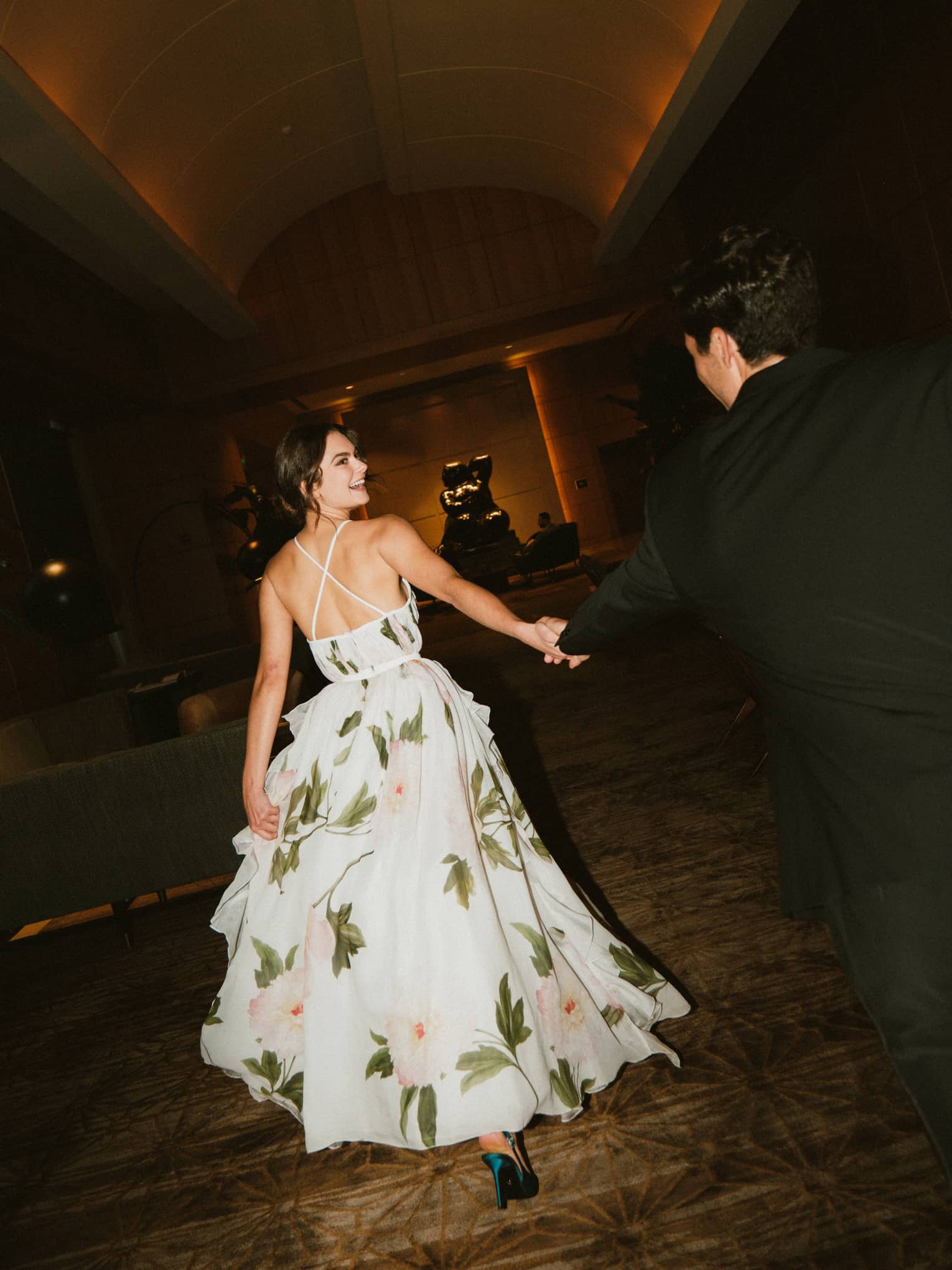Couple dressed in formalwear walks hand in hand along a dimly lit corridor