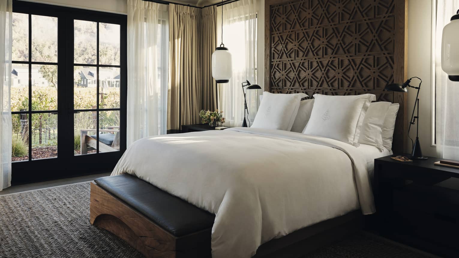 Luxury residential villa bedroom with a king bed, carved wood headboard, bench, nightstands, pendant lights and large windows with a vineyard view