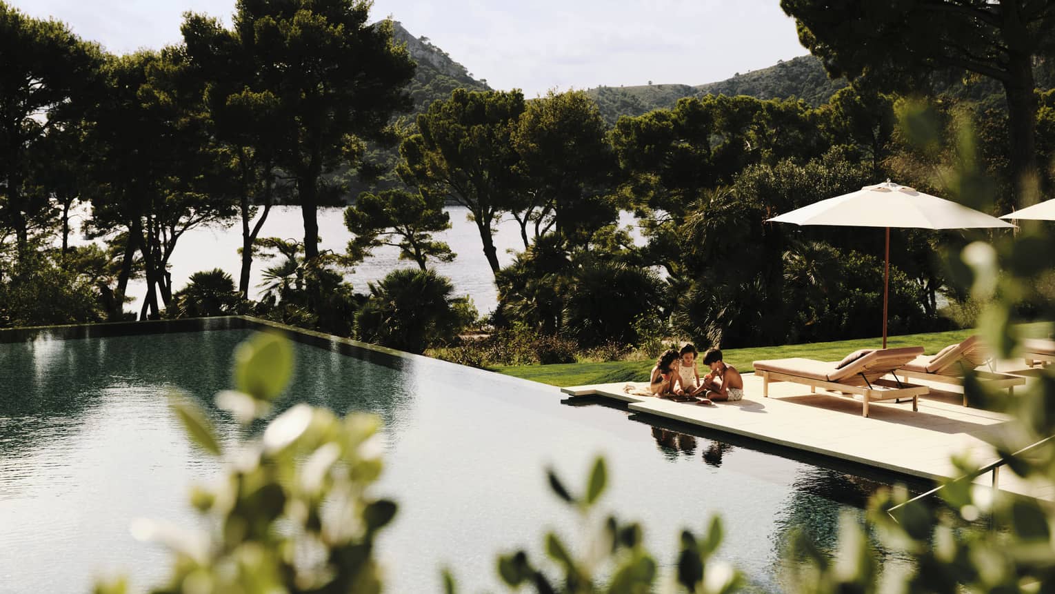 young children play on pool deck next to large resort pool, surrounded by push pine trees