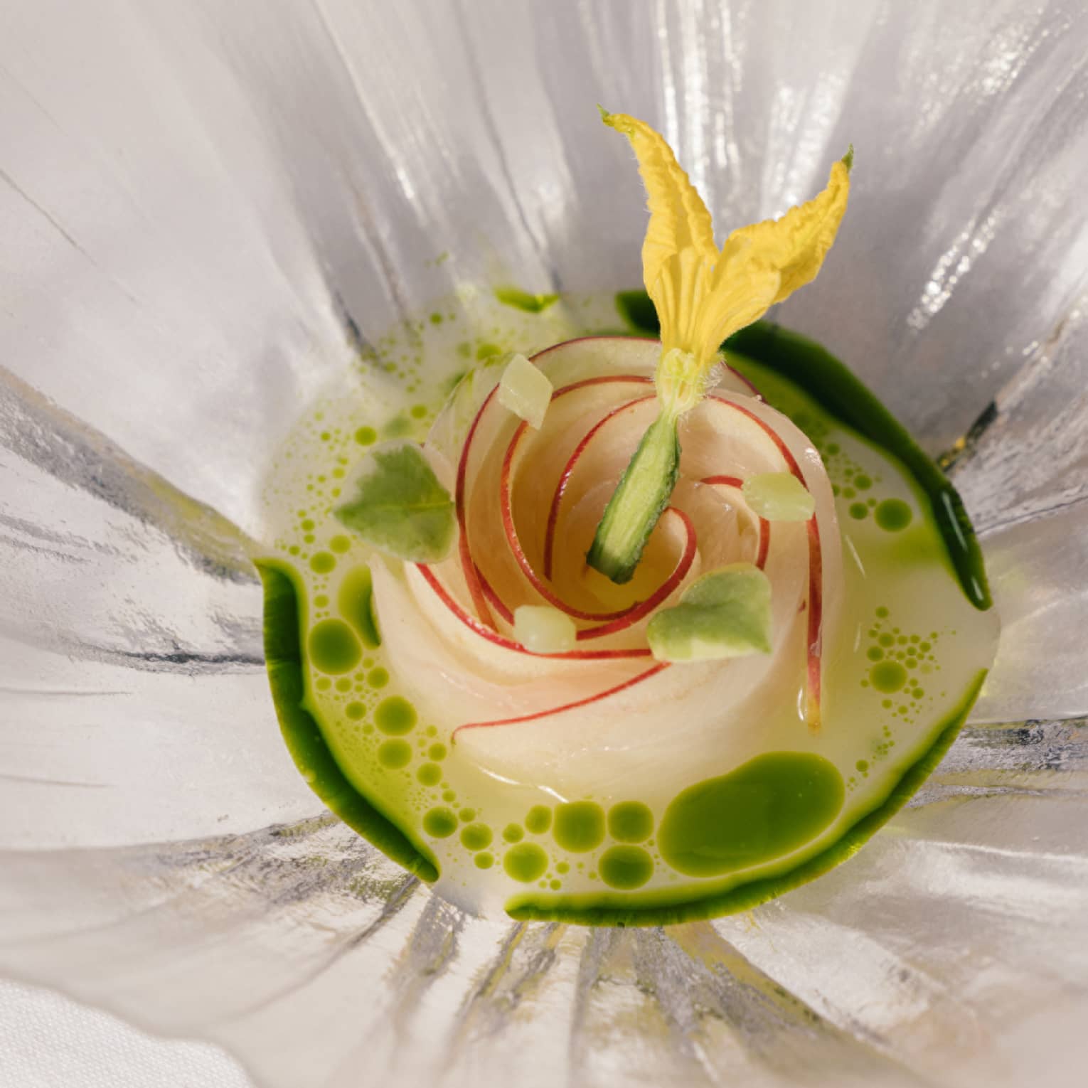 Elegant dish arranged as a rose, topped with a yellow edible flower, in a green herb-infused broth