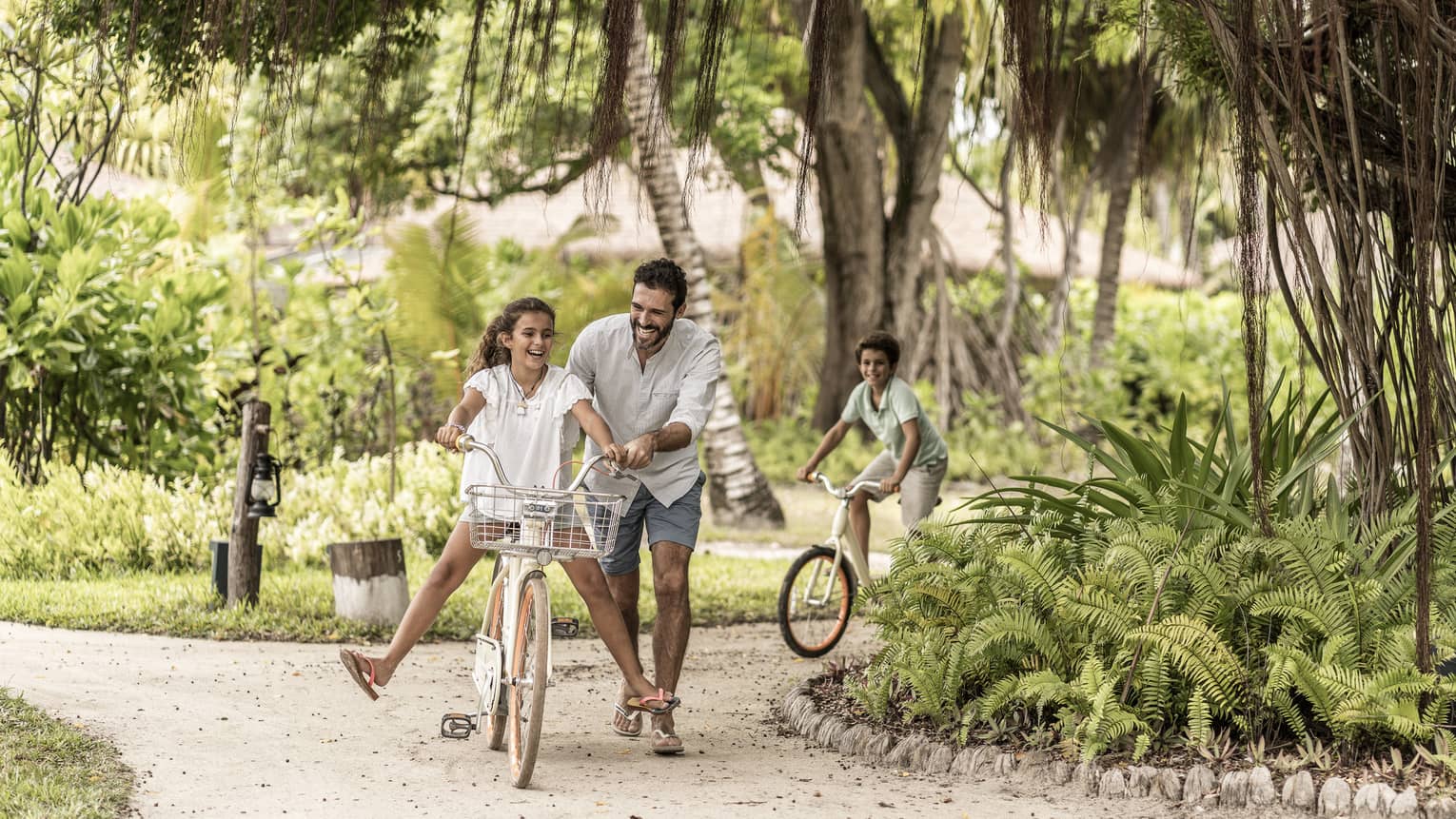 Girl riding bike, laughing with legs off pedals, dad beside holding on and brother on bike behind