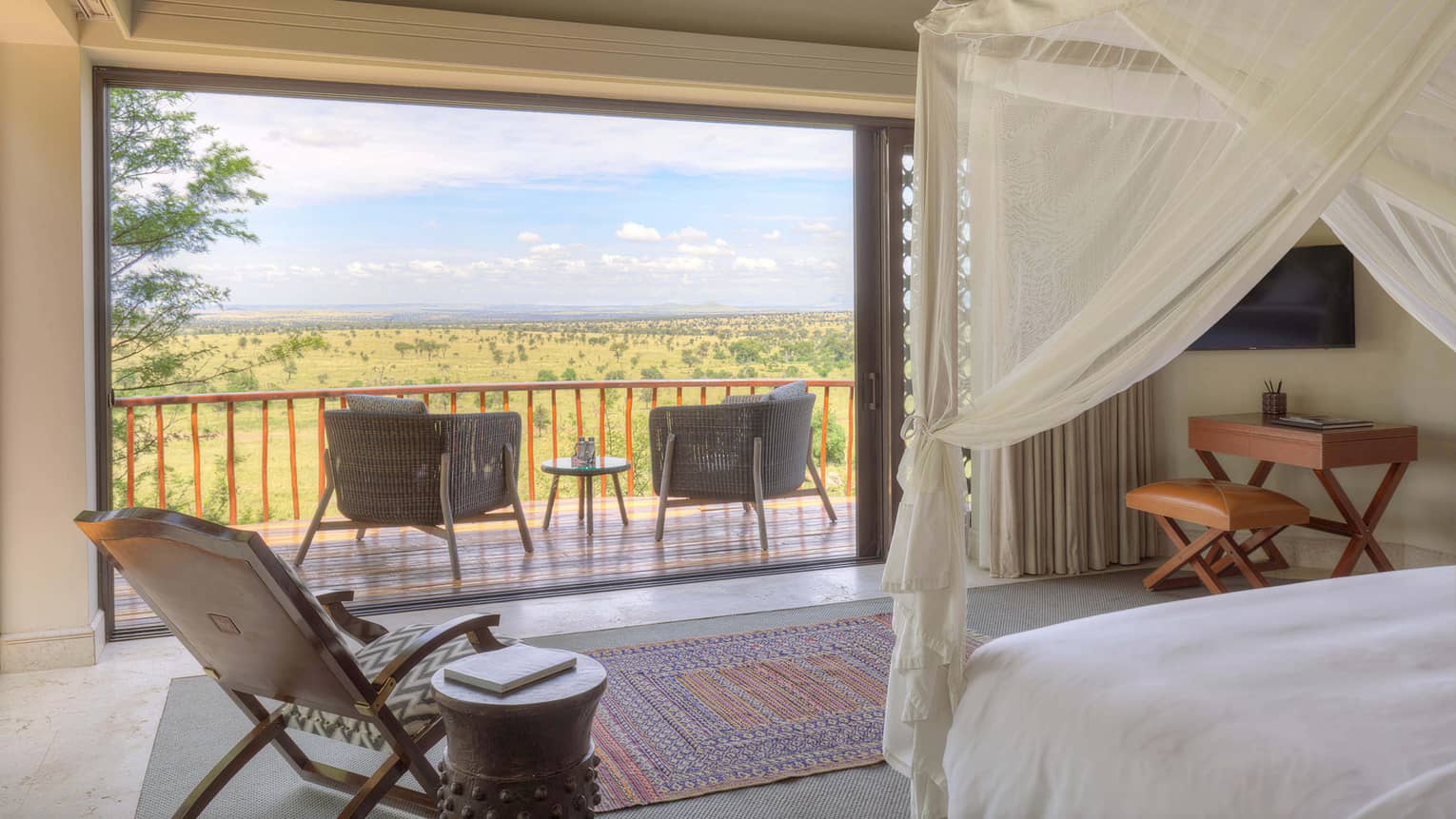 Villa bedroom with airy fabric-draped bed, side chair with round table and desk facing deck with chairs looking out to expansive plain
