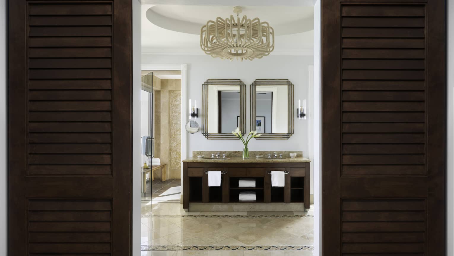 Elegant beachfront villa residence bathroom with double vanity, framed mirrors, and a chandelier, viewed through dark wooden doors