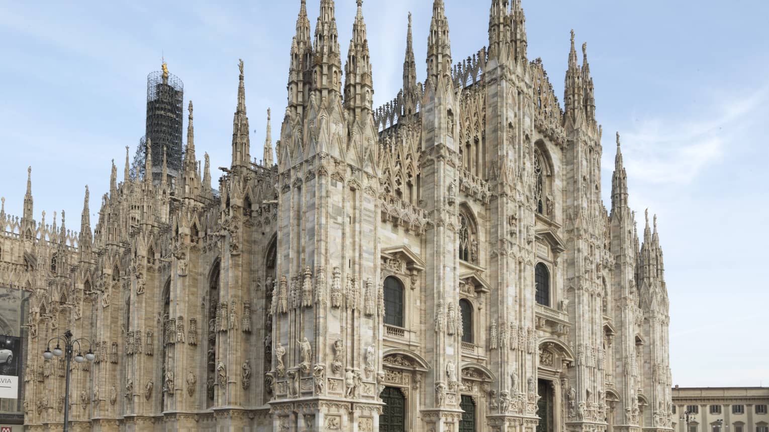The exterior of the Milan Cathedral (Duomo di Milano), showcasing its intricate Gothic architecture with tall spires and detailed stone carvings against a clear sky.