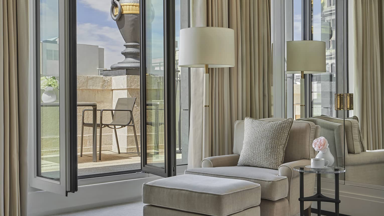 Luxury hotel suite with beige armchair, matching ottoman, floor lamp, glass side table and open balcony door overlooking terrace with chair and stone railing