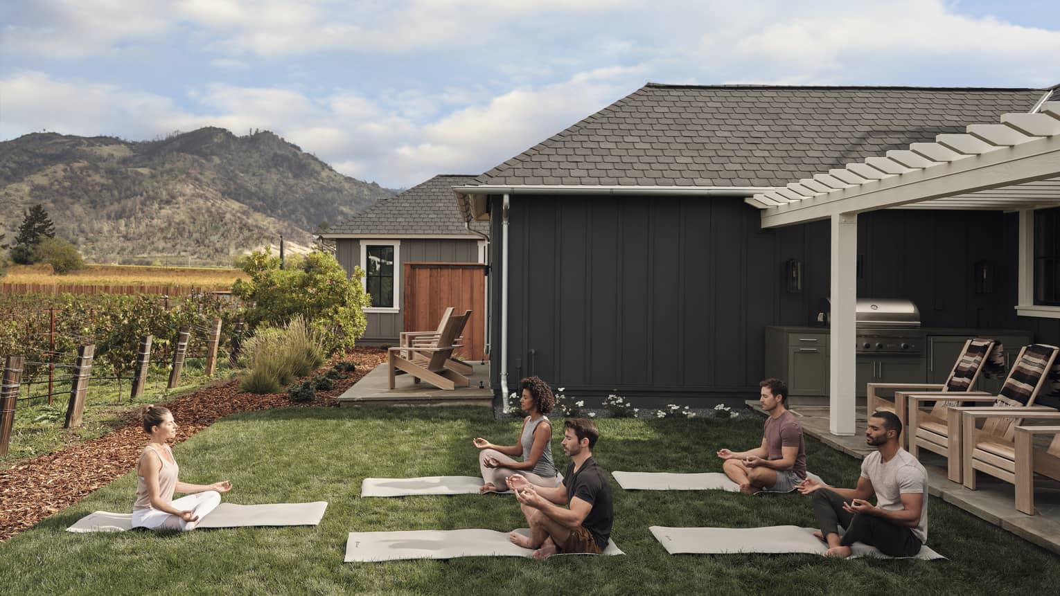 Group of people practicing yoga on mats in a grassy villa backyard near vineyards, with mountain views, wooden chairs and an outdoor grill area under a pergola