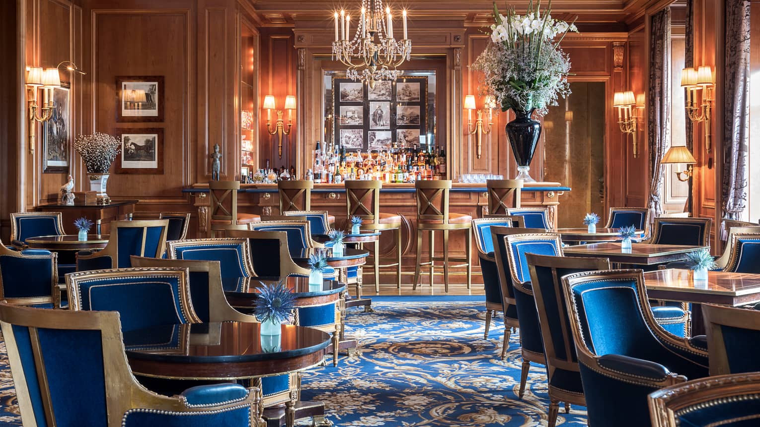Le Bar des Bergues lounge with elegant blue velvet chairs with wood trims around cocktail tables, crystal chandelier, bar