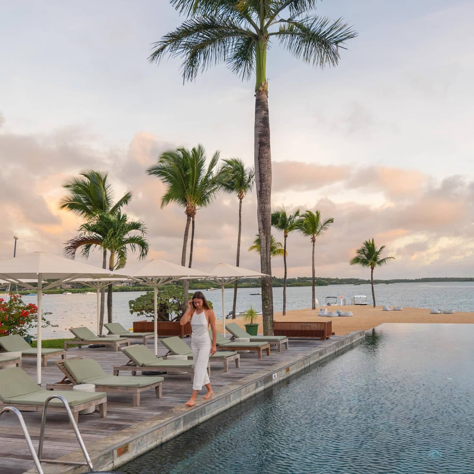 A woman walks by a row of lounge chairs at an oceanside pool at sunset