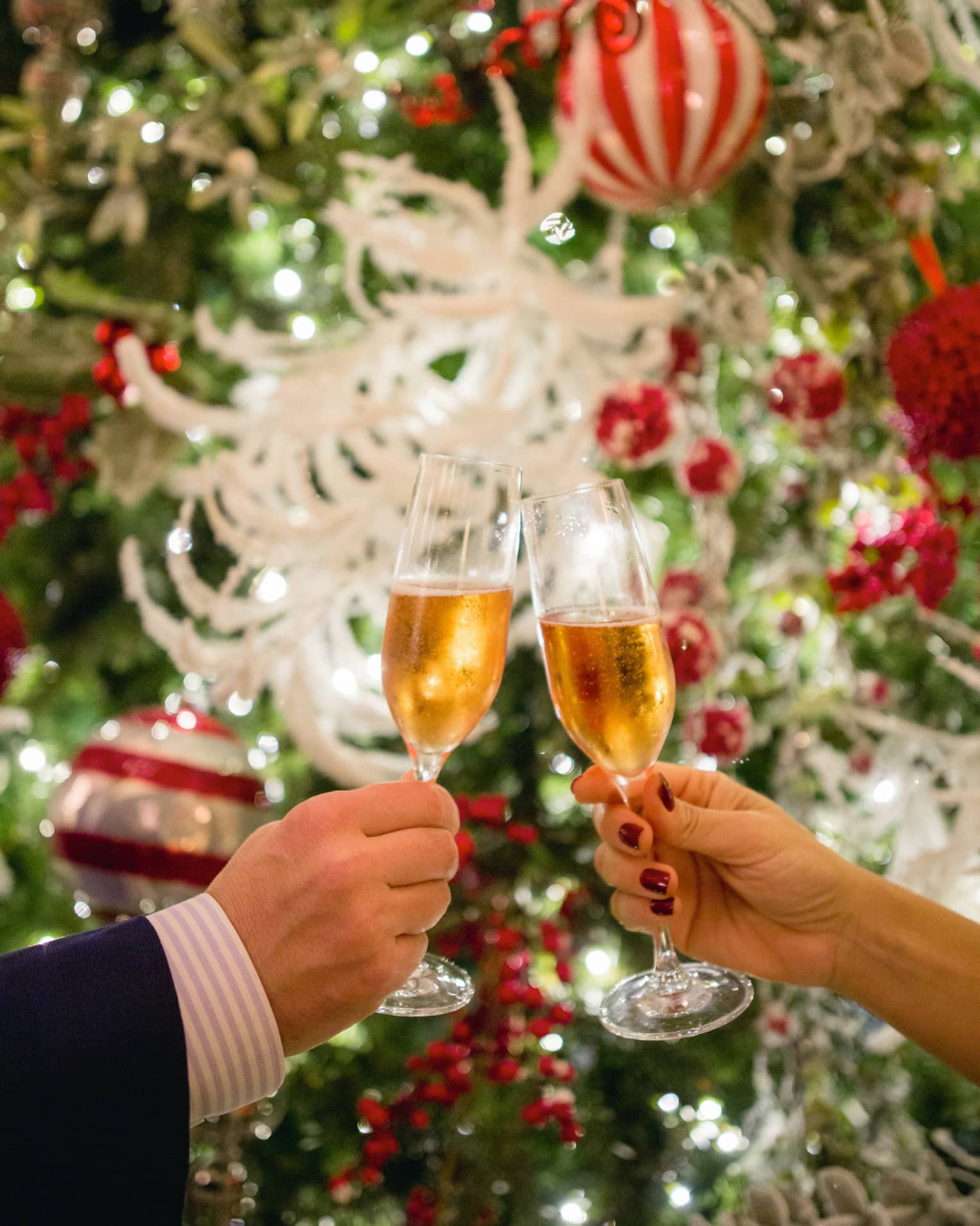Two hands toasting Champagne glasses with holiday decor in backdrop
