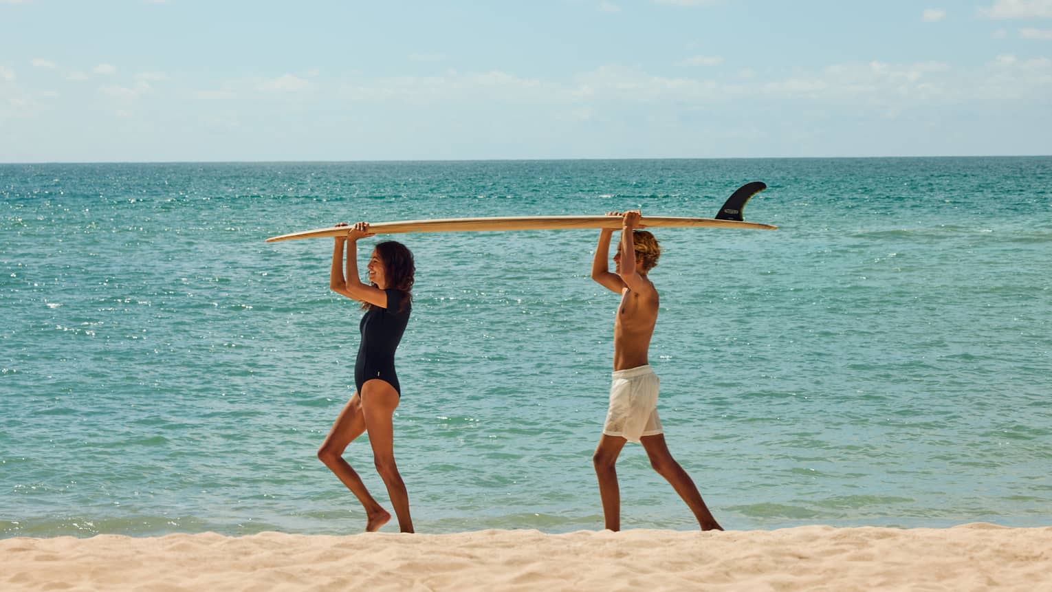 Two kids walk on beach with a surfboard on their head