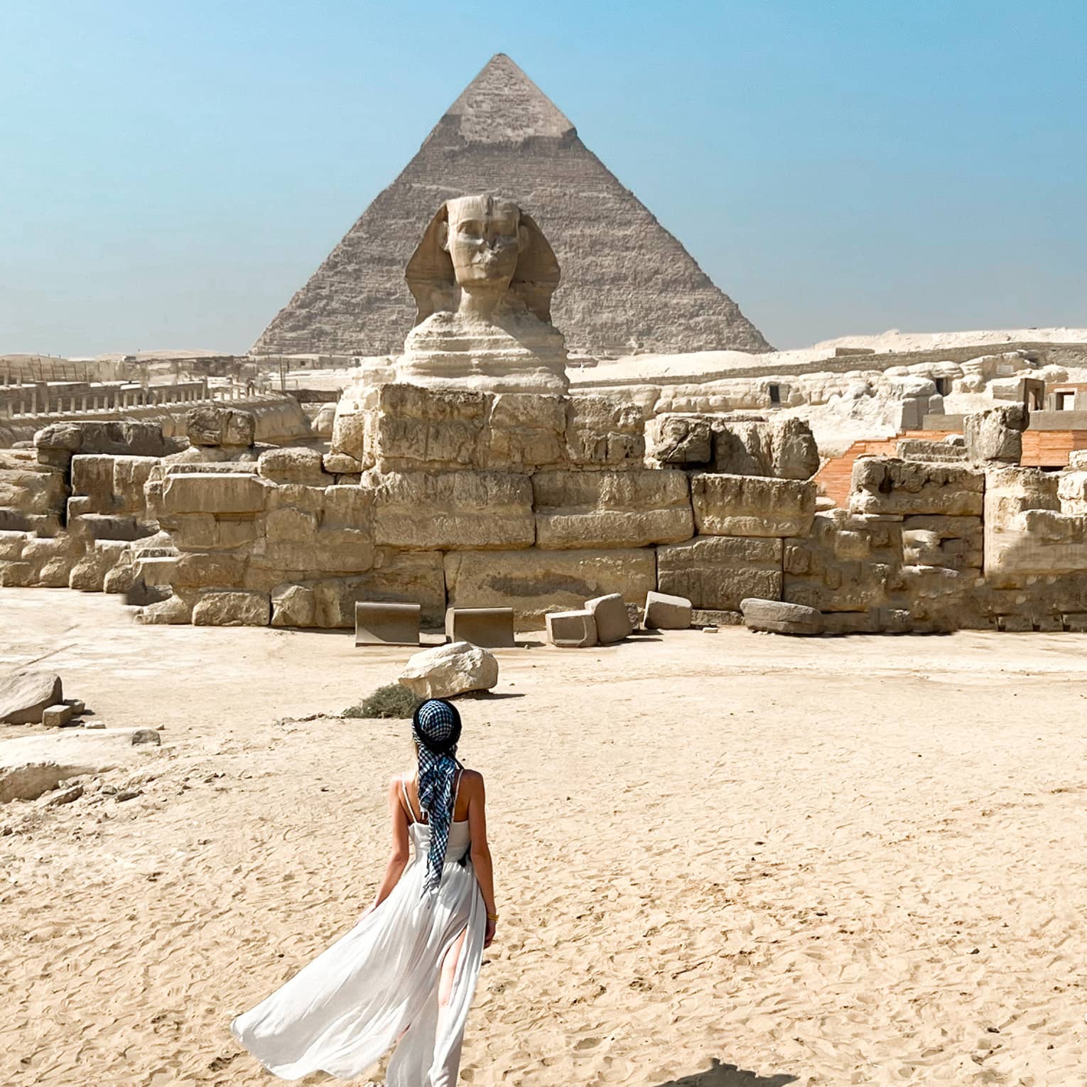 View from behind as a guest crosses golden desert sand toward the immense ruins and sphinx, a pyramid rising up beyond.
