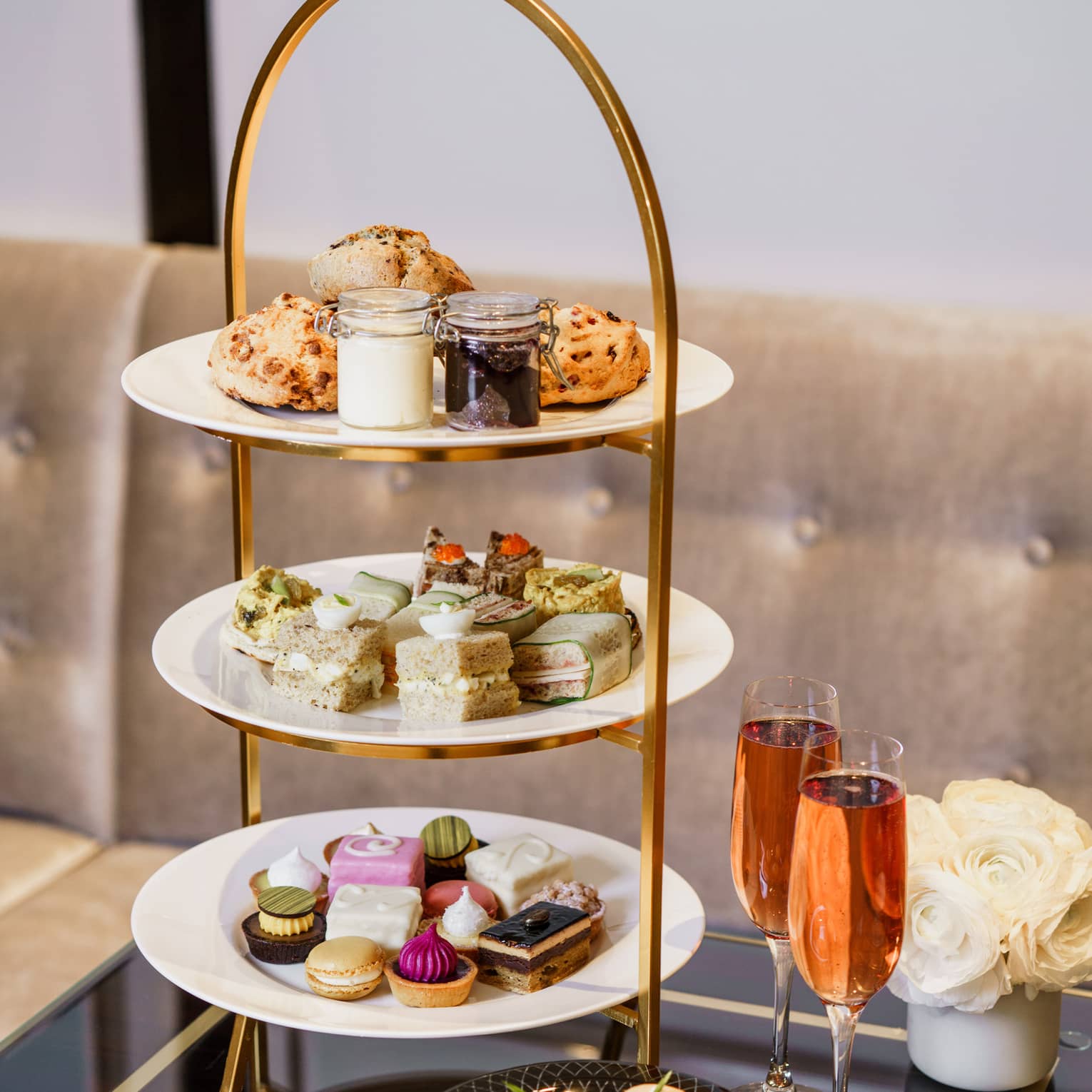 A tiered tray of finger sandwiches, colourful desserts, pastries, and cream and jams, alongside two flutes of sparkling wine.