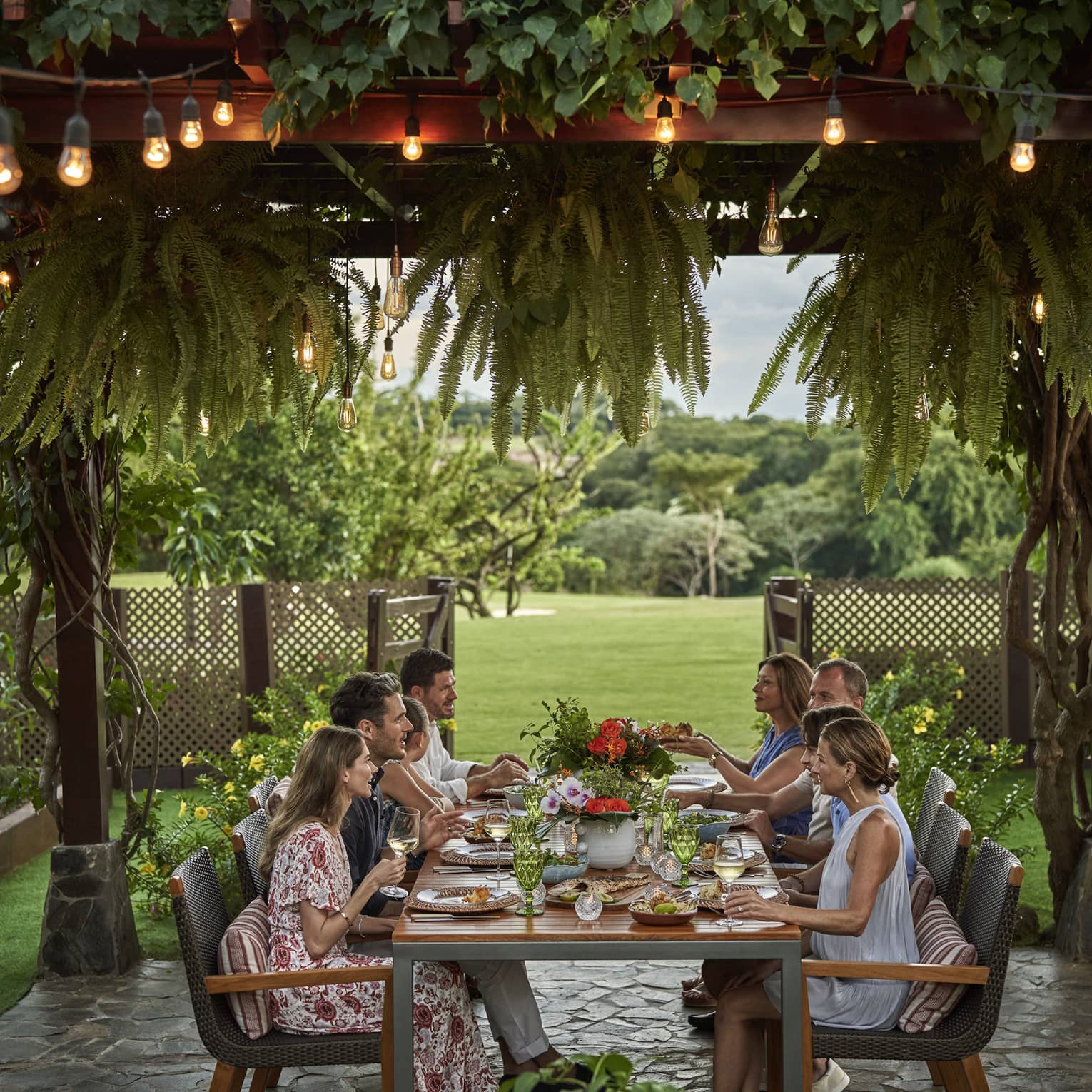 Eight people sit around a long dining table set up outside beneath a canopy of greenery and string lights
