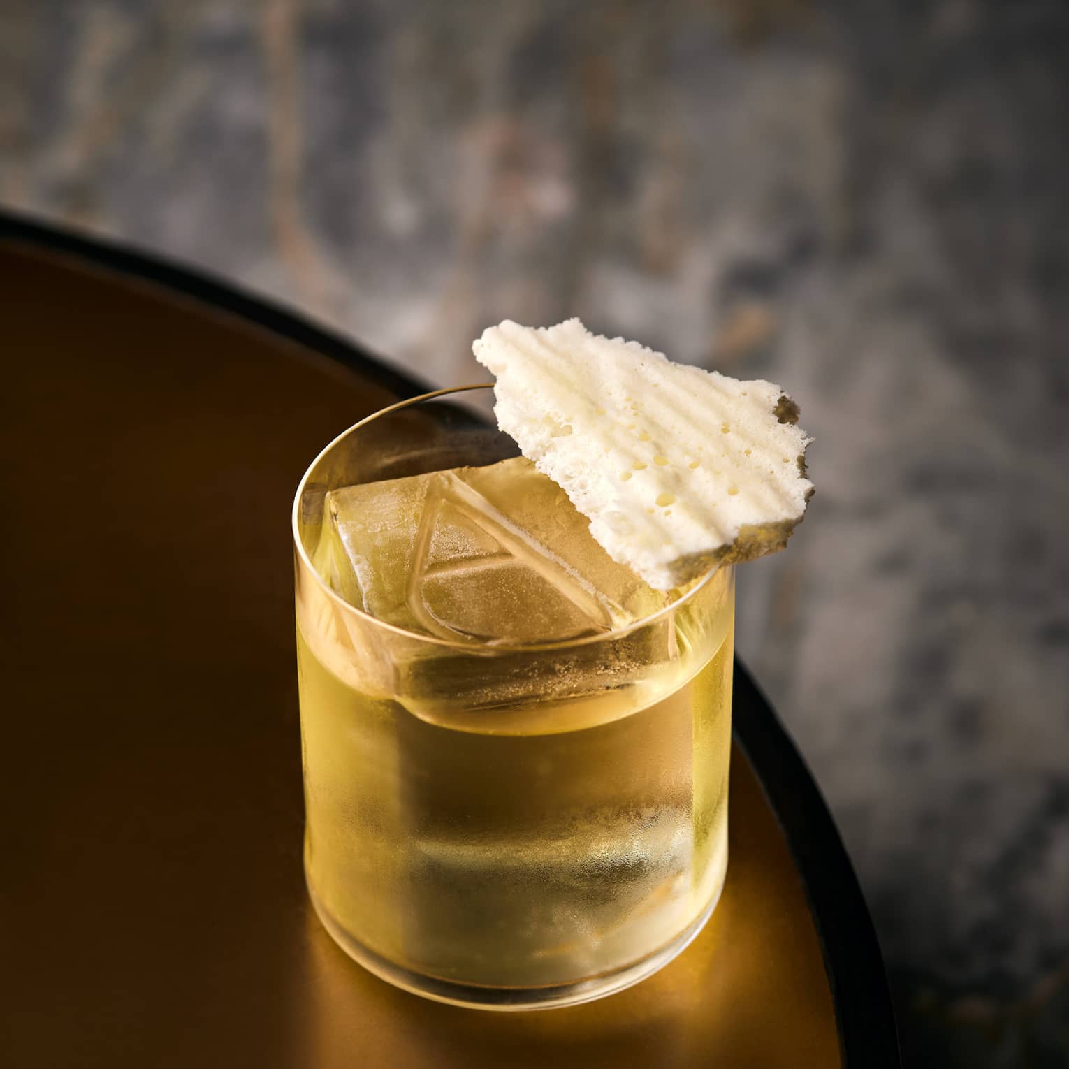 Yellow cocktail in a low glass with a large cube of ice, engraved with the letter A