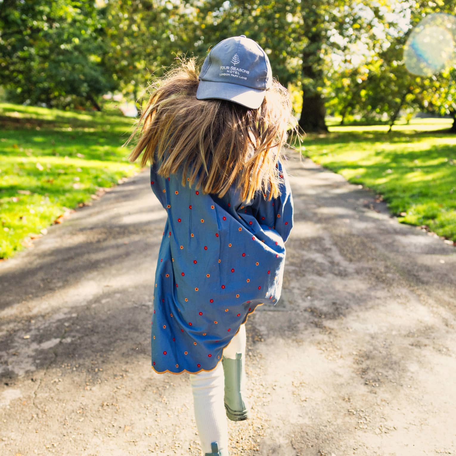 Rear view of long-haired child on a sunny path flanked by green grass and trees, wearing backwards London Park Lane ball cap.