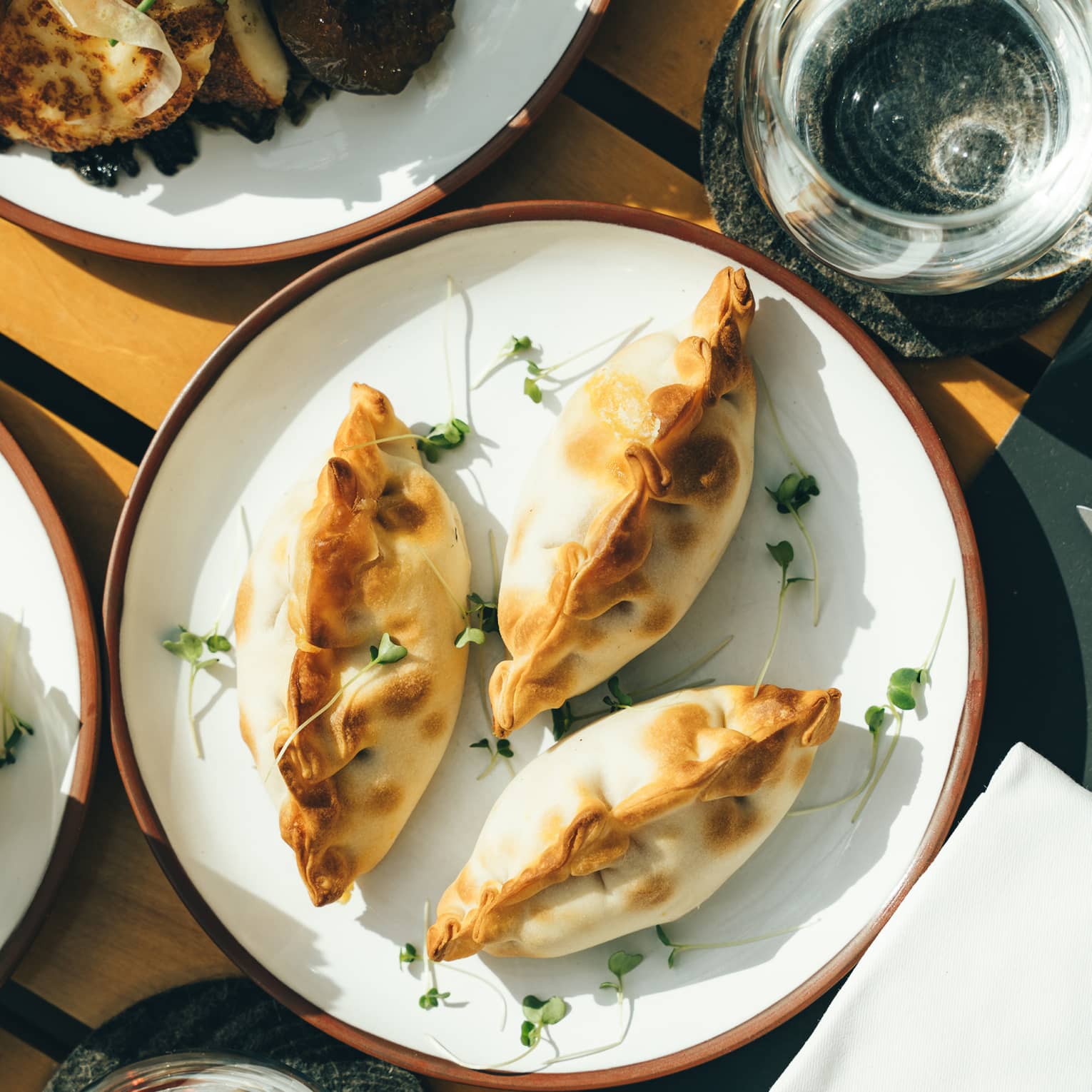Three plump, golden-brown empanadas on a porcelain dish surrounded by sunlit water glasses and other plates of fine foods.