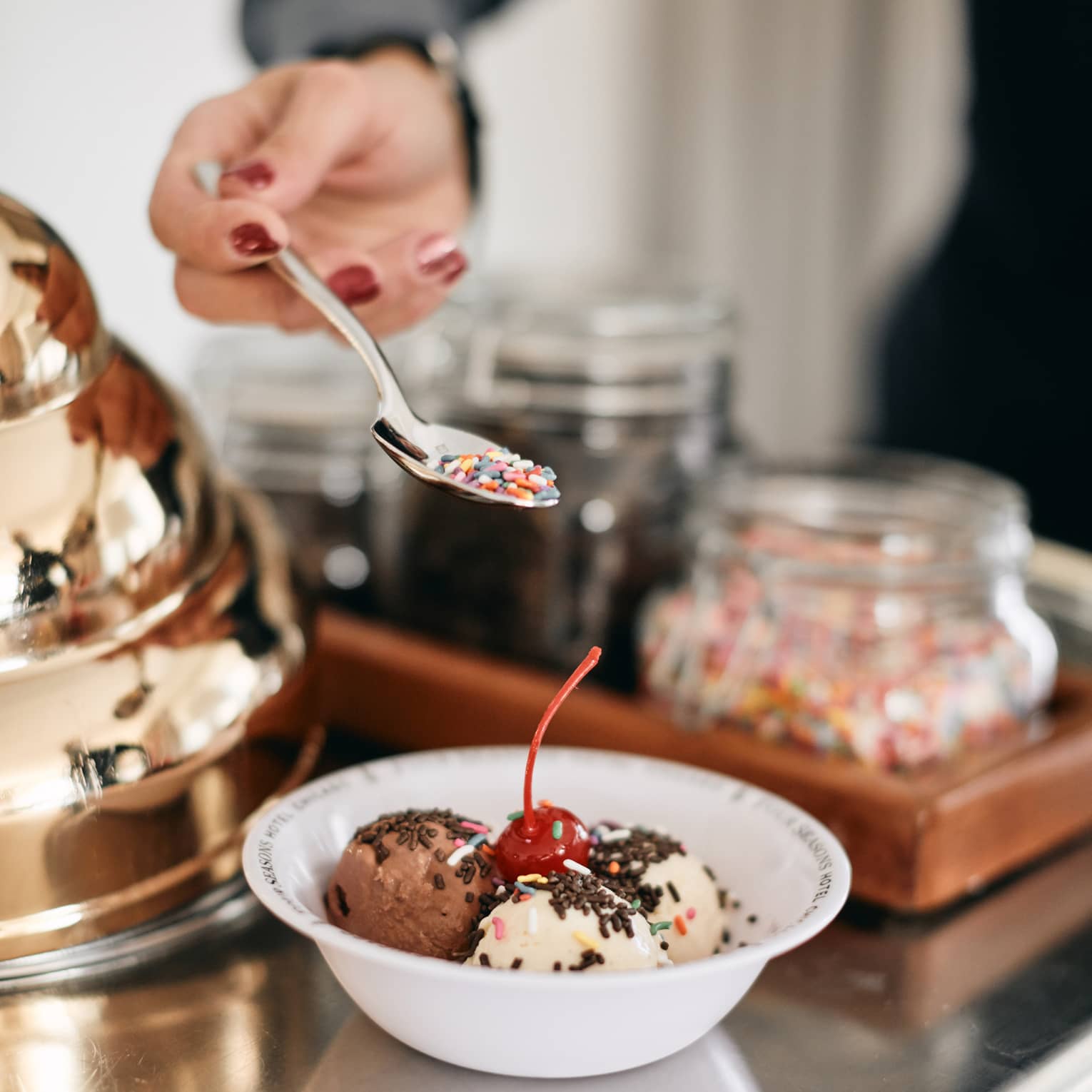 A member of staff creating a colorful ice cream bowl for a guest, there are various flavours in the bowl, sprinkles, and a cherry.