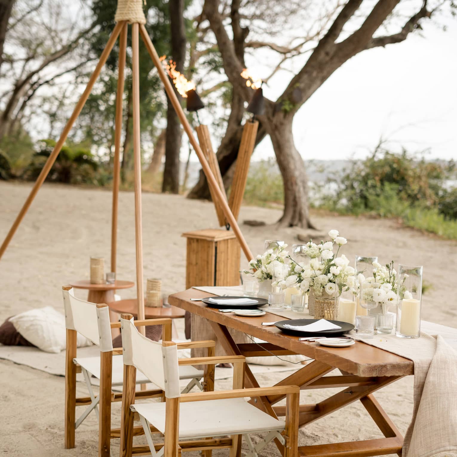 Wooden dining table and chairs set up on a beach, decorated with white floral centrepieces and candles