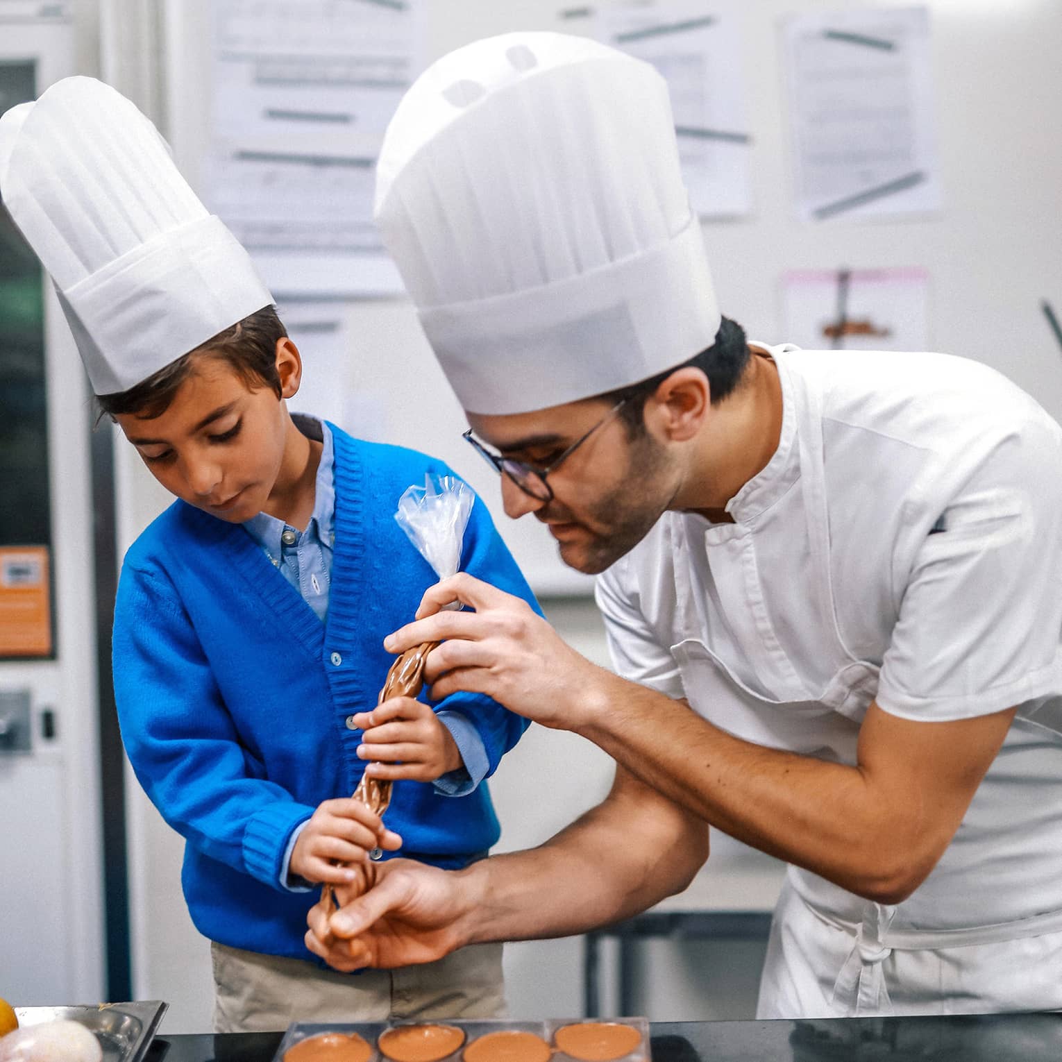 Chef helping a young child pipe chocolate into molds during a cooking class, both wearing white chef hats.
