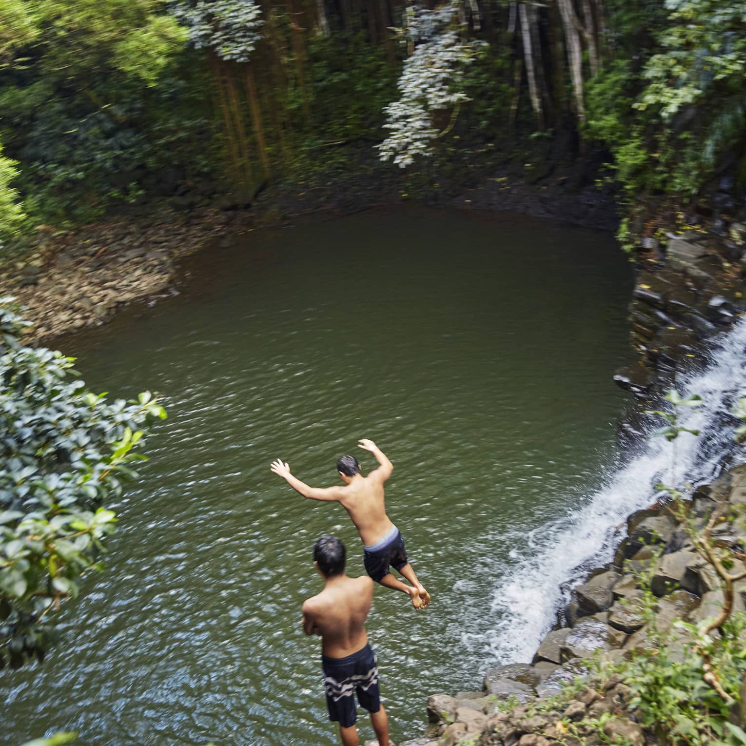 Two people leap from rocks to water below waterfall on the road to Hana