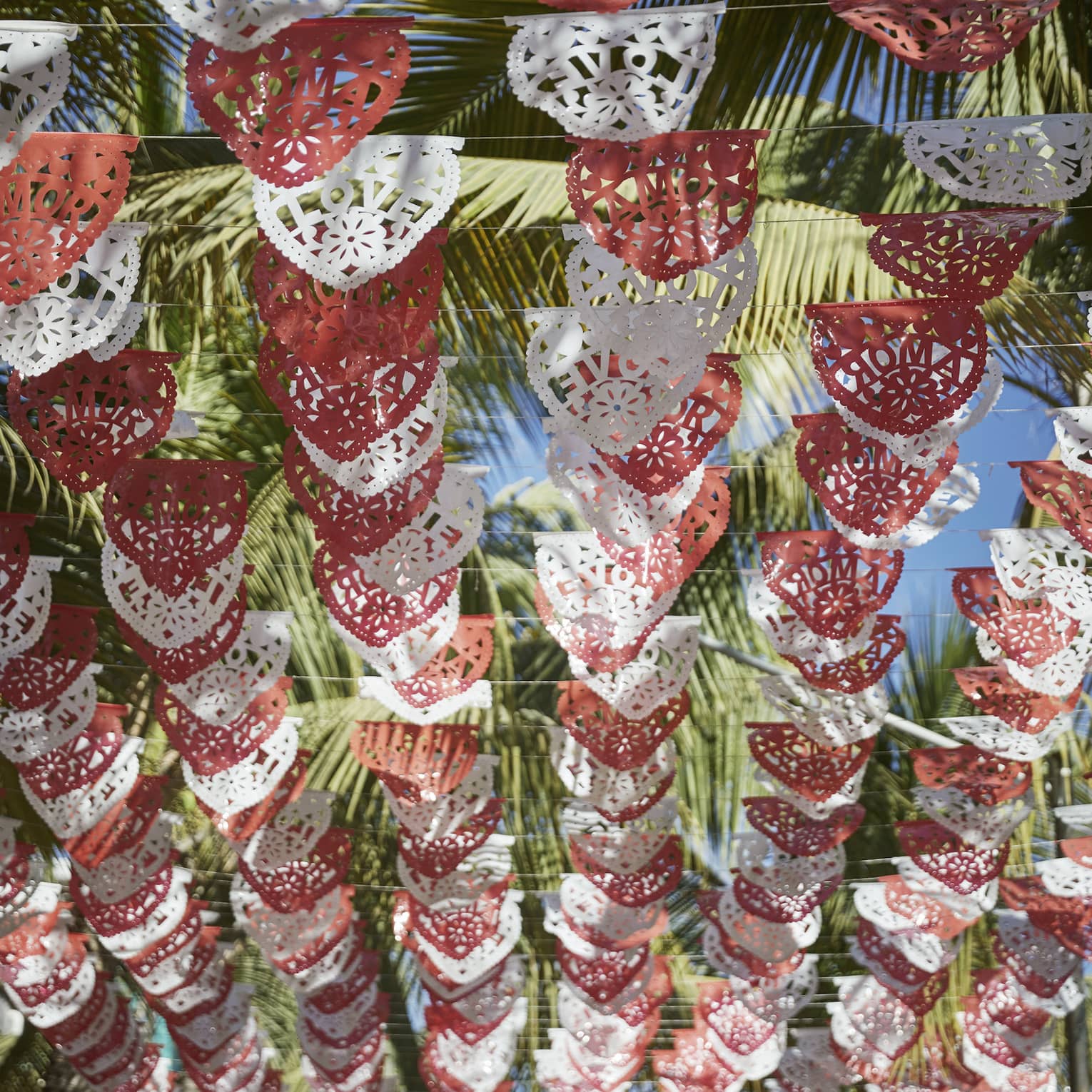 Looking up at rows of red and white Mexican Papel Picado banners hanging above, with palm trees and a sunny sky above them.