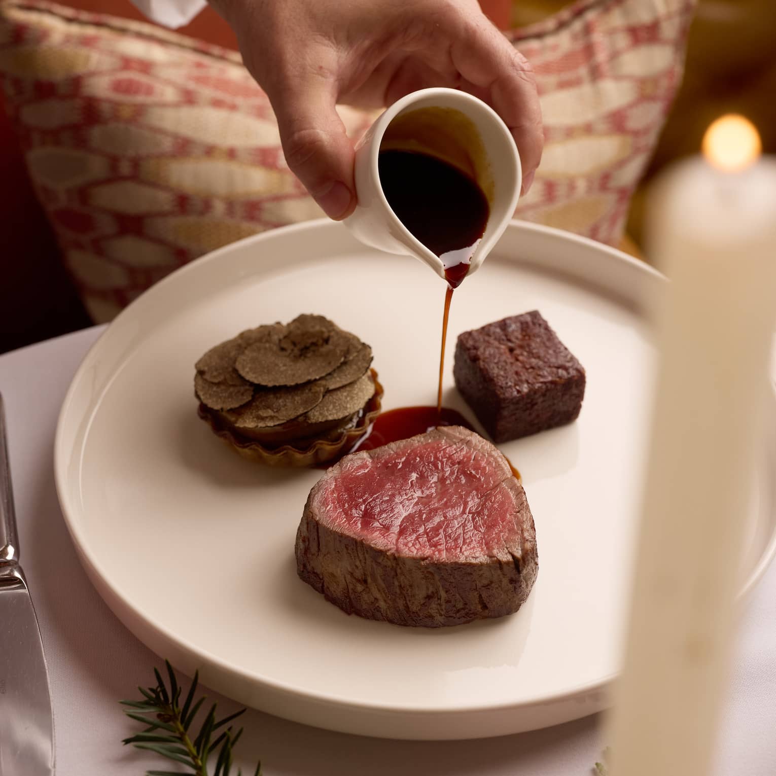 A person pours sauce on a plate of food items, including a slice of cooked meat, set on a table with holiday decor.