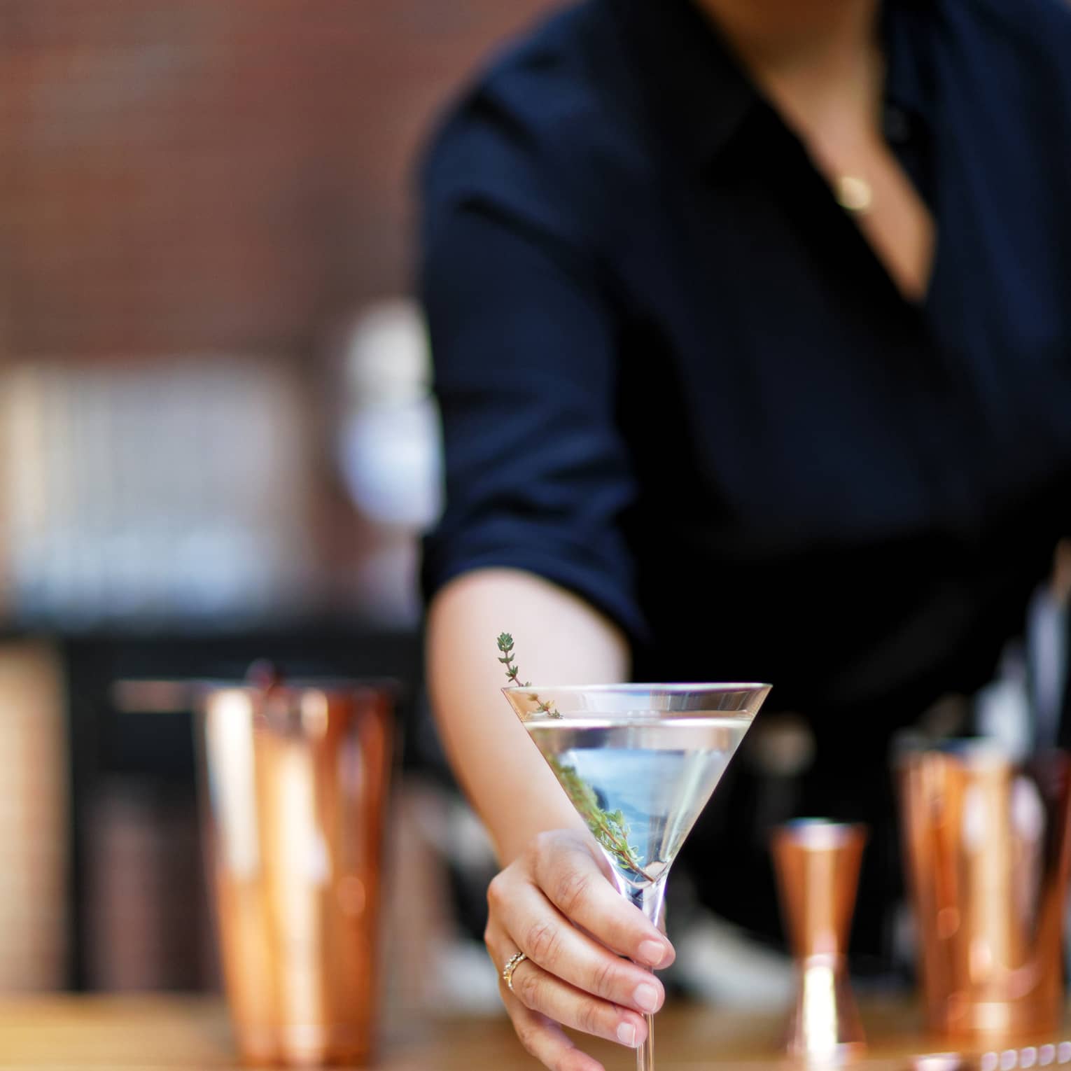Bartender offering a martini garnished with a sprig of fresh thyme set on a small napkin, cocktail shakers in the background.