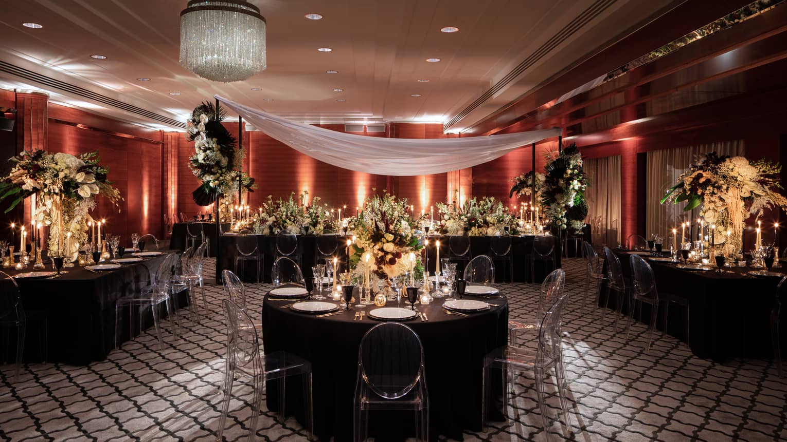 Elegant wedding reception setup at the Four Seasons London Park Lane in Mayfair, featuring a grand ballroom with rich red and gold decor. The room is arranged with round tables draped in black linens, each adorned with tall, opulent floral centerpieces featuring white and gold flowers. Crystal chandeliers and sophisticated lighting cast a warm glow over the scene, enhancing the luxurious atmosphere. A sheer white fabric swathes gracefully across the ceiling, adding a soft, romantic touch to the sophisticated and festive setting.