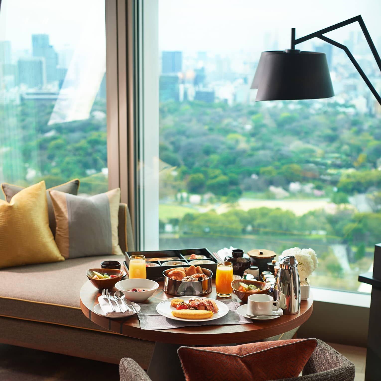 A breakfast tray of pastries, fresh fruit, juice and coffee on a guest room lounge chair at a window overlooking lush gardens.