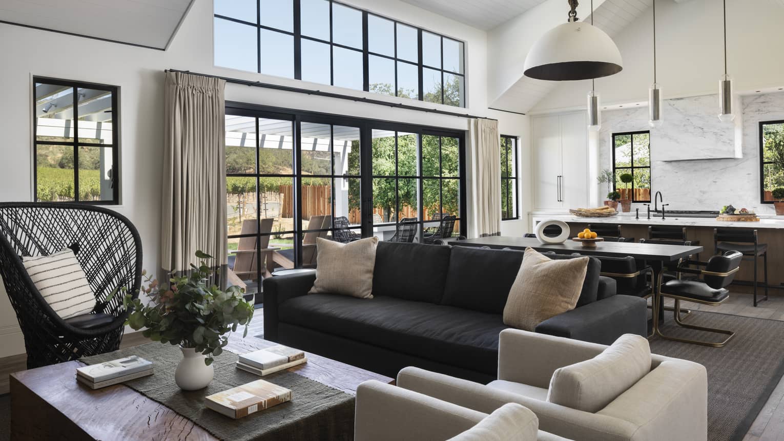 Open-concept living and kitchen area with a black sofa, beige armchairs, wicker chair, dining table, marble island and windows with views of the outdoor patio