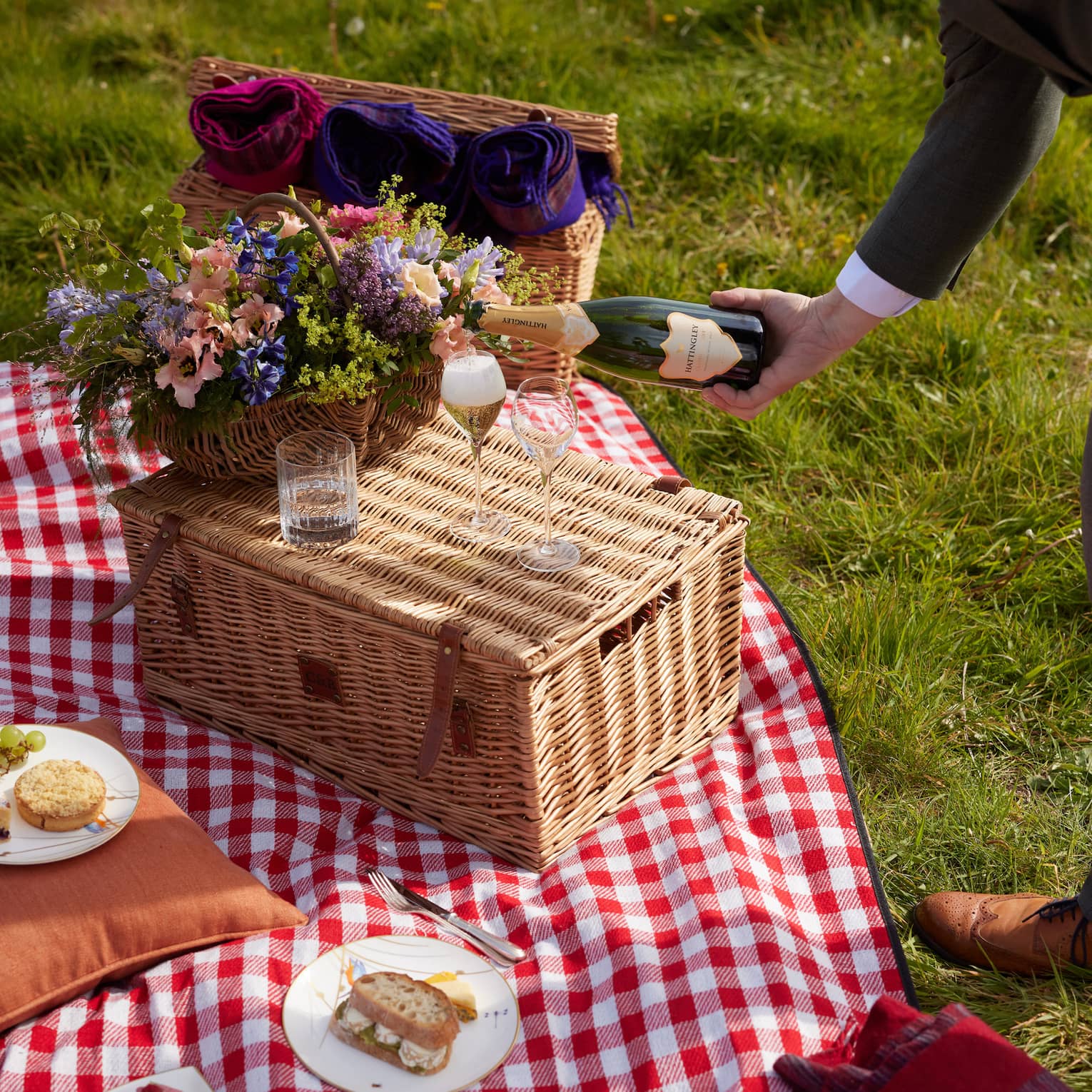 Attendant pours champagne into glass beside bouquet atop wicker picnic basket on a red-and-white gingham blanket