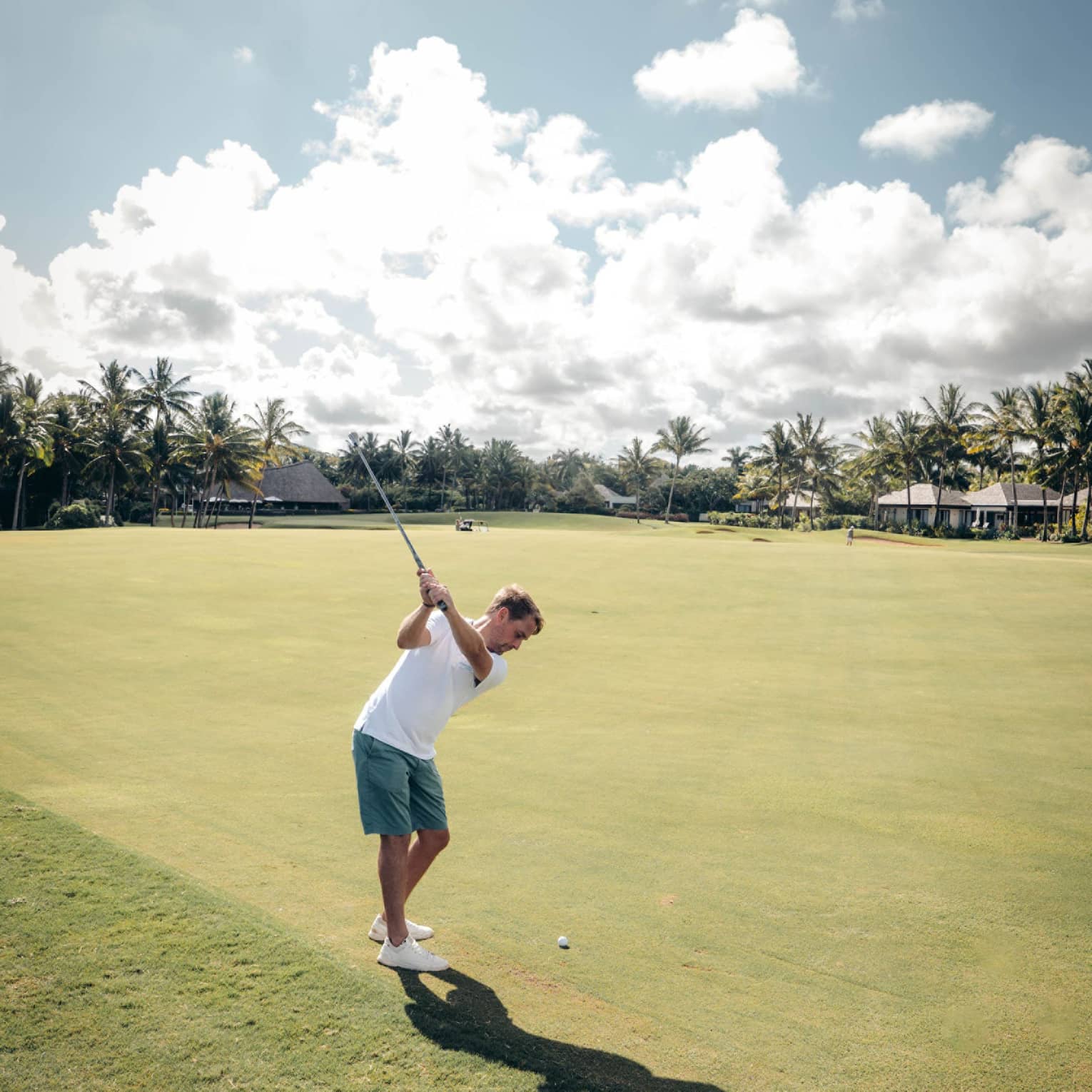 Man in white polo and green shorts swinging a golf club on green with palm trees and cloud-dotted sky in backdrop