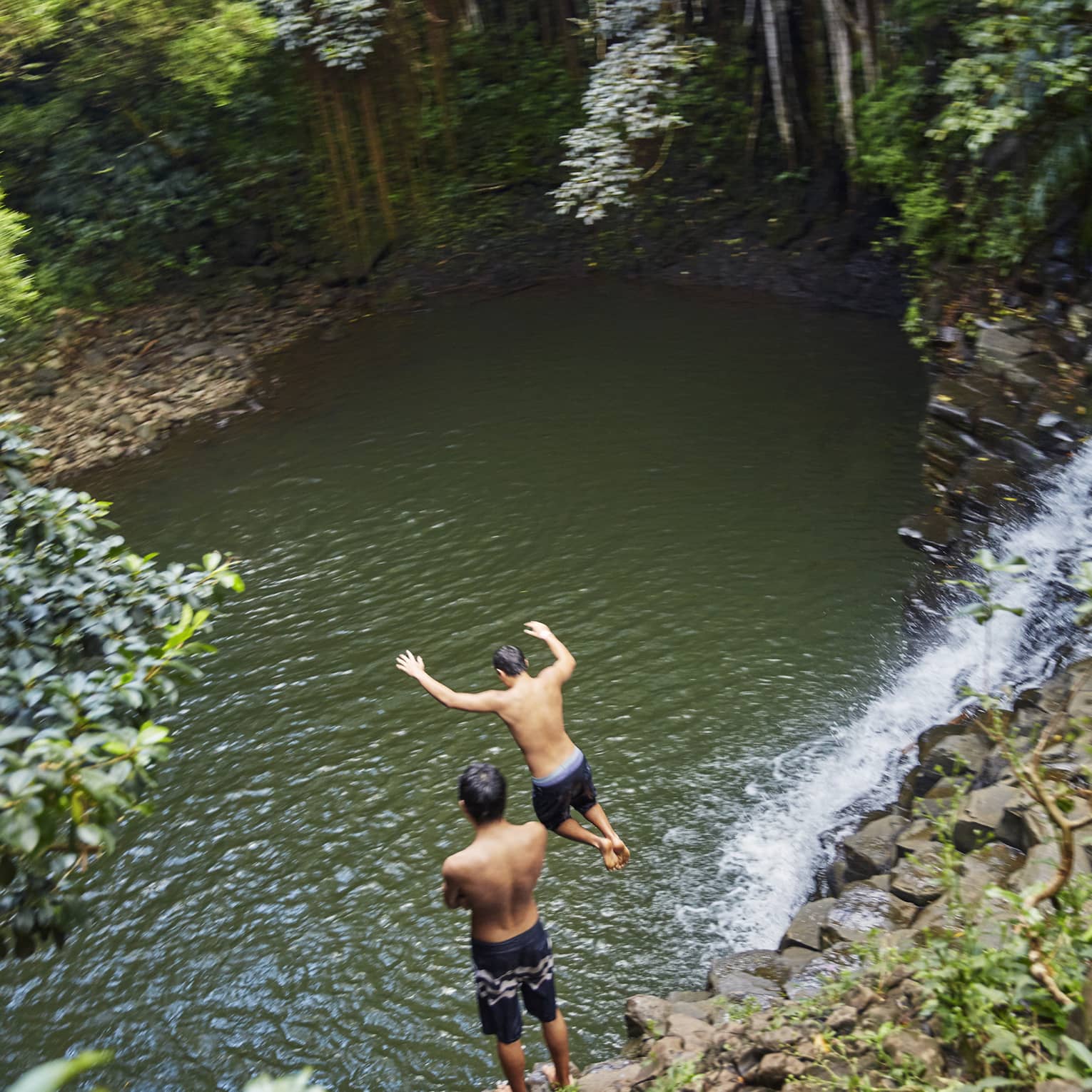 Two people leap from rocks to water below waterfall on the road to Hana