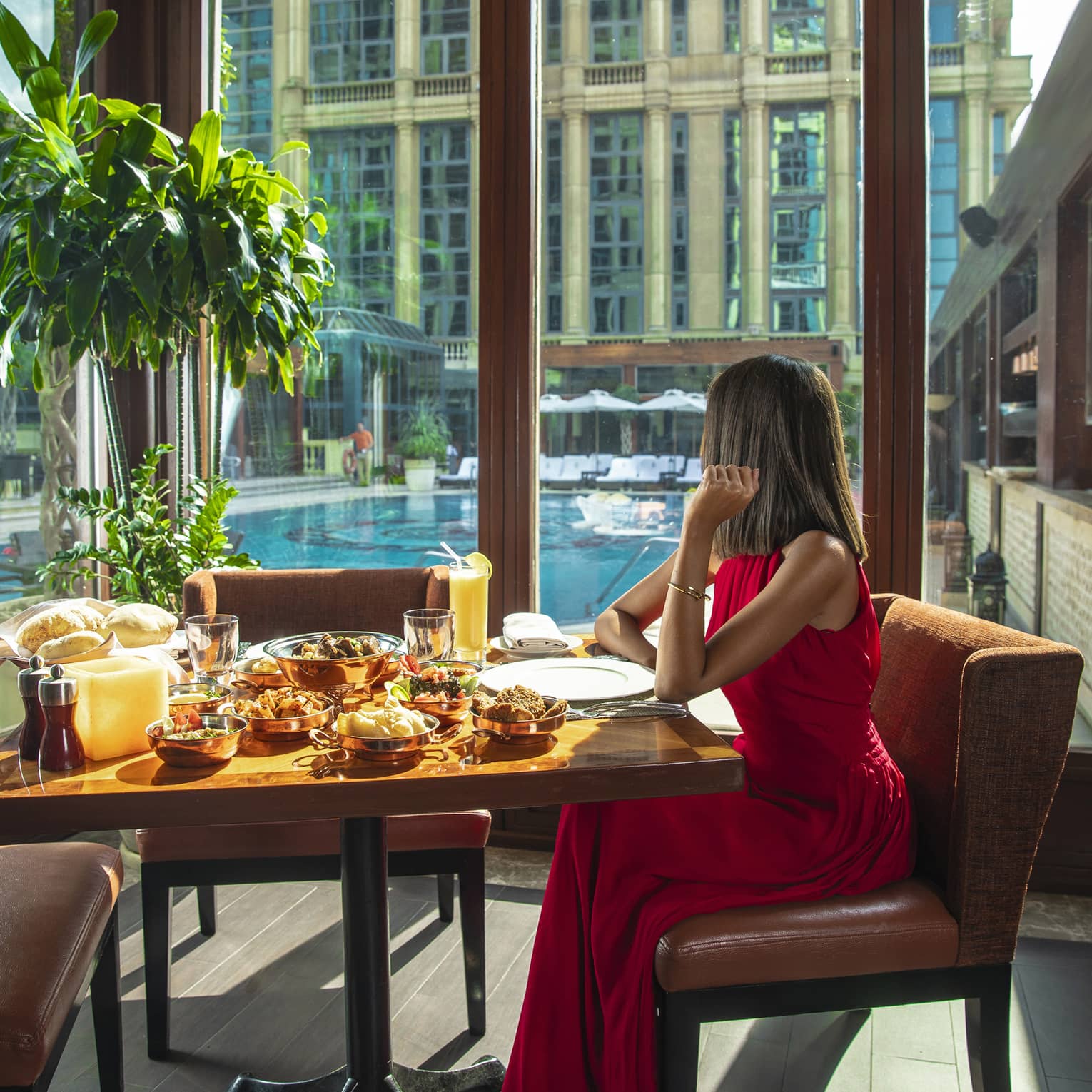 A woman in a long red dress sits at a table with a variety of food next to a long window overlooking a pool