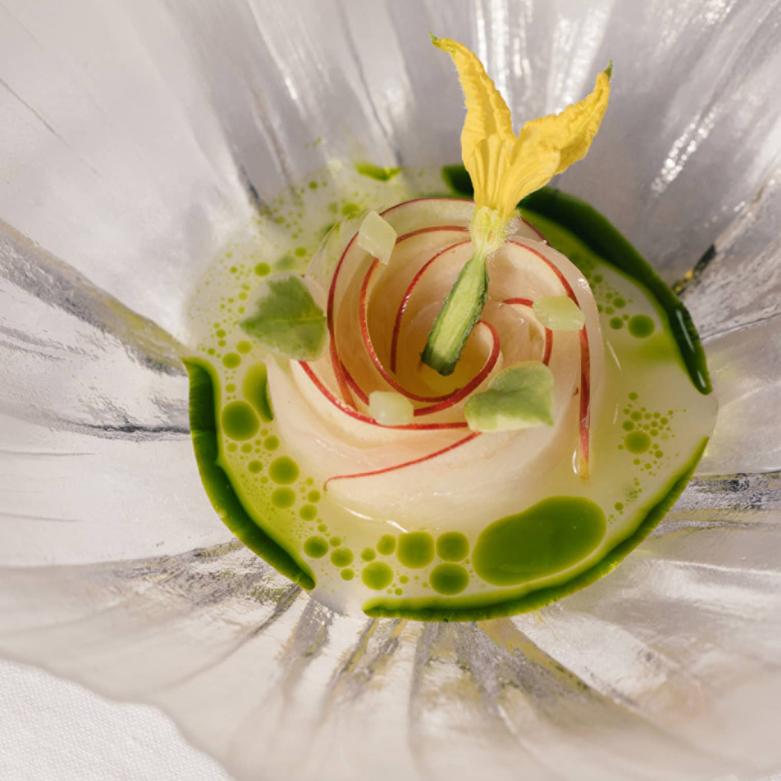 Elegant dish arranged as a rose, topped with a yellow edible flower, in a green herb-infused broth