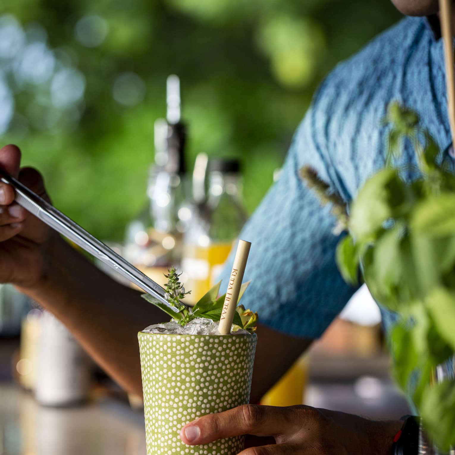 A smiling bartender in a blue shirt garnishes a cocktail; blurred bottles of liquor and green foliage in the background.