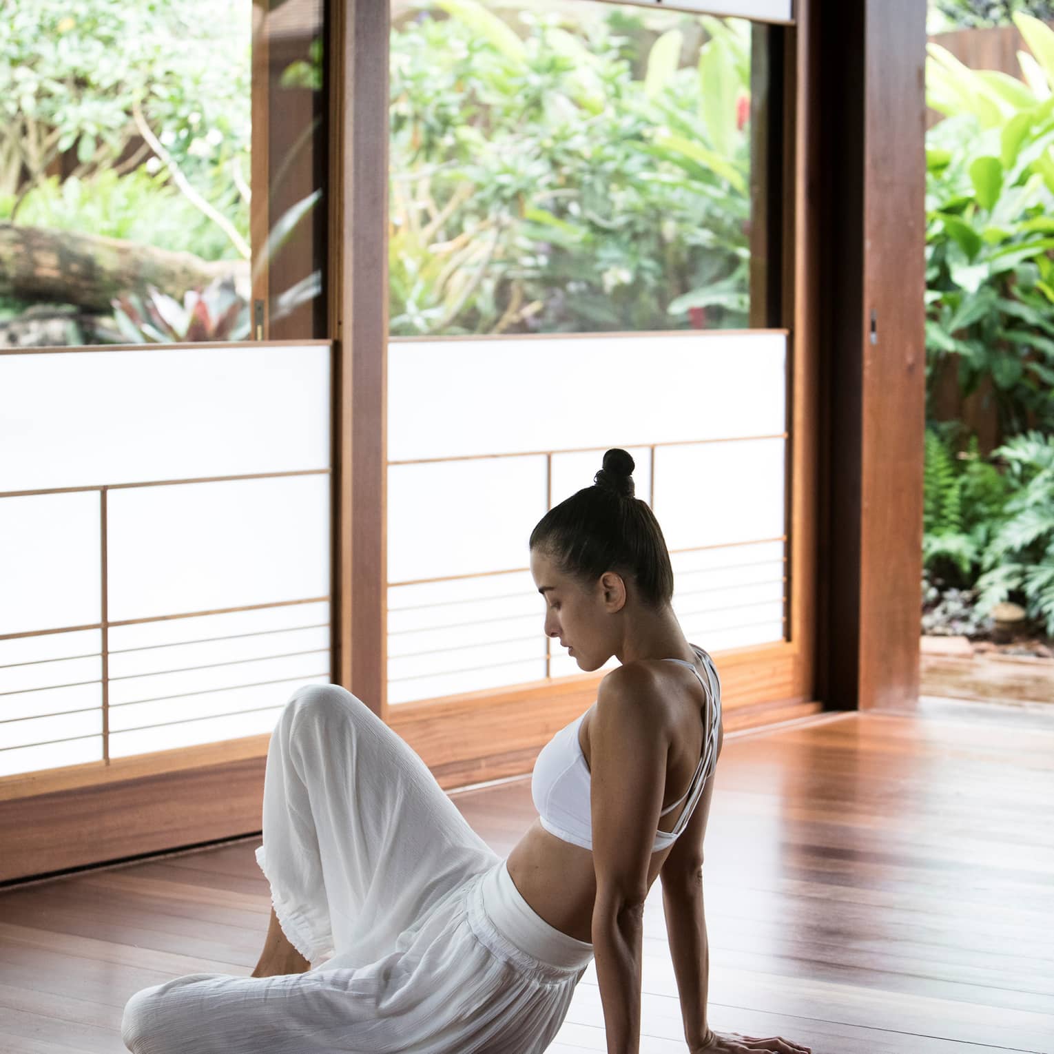 A woman dressed in workout clothes sits on the floor of a room with wooden walls and windows overlooking lush greenery